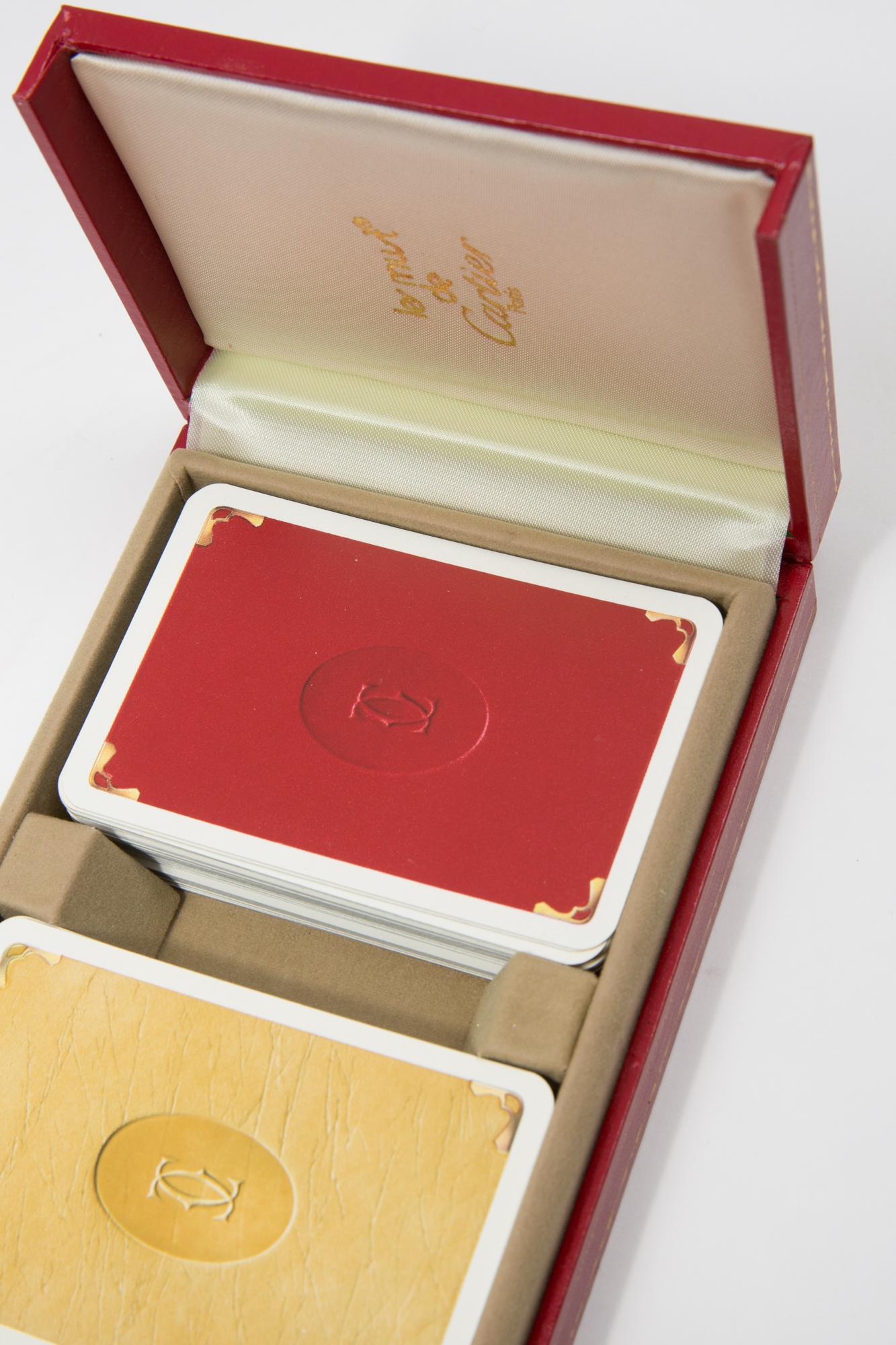 Coffret de cartes à jouer Cartier comprenant un jeu plus un joker, des cartes de poker et de bridge dans un étui rouge à 2 portes de Cartier. Le coffret de cartes doublé de soie, aux coins dorés 18 carats, imprimé du logo Les Must de Cartier, elles