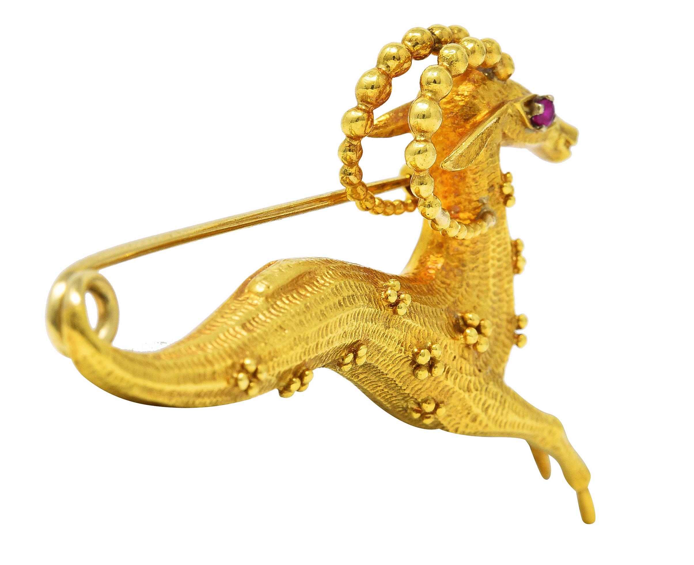 Brooch is designed as a stylized leaping gazelle with grooved textured body. Featuring notched curling horns and gold granulation throughout. Accented by a prong set round cut ruby eye - transparent medium red in color. Rear of body curls into