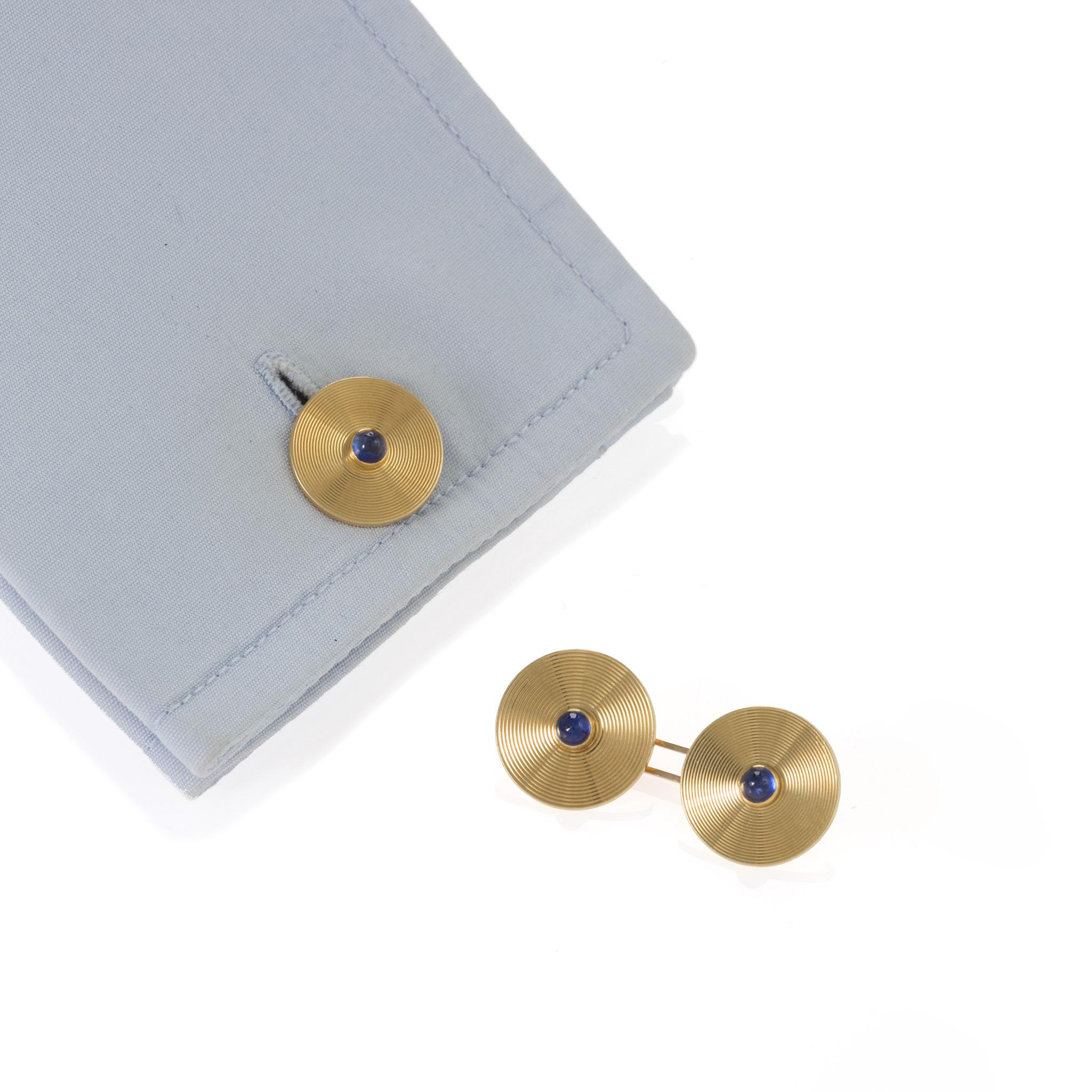 Dating from the Art Deco period, these circular gold cuff links are set with sapphire cabochons. Designed as double links, each engine-turned disk centers a bezel-set sapphire cabochon, combining the bright blue of the sapphires with rich yellow