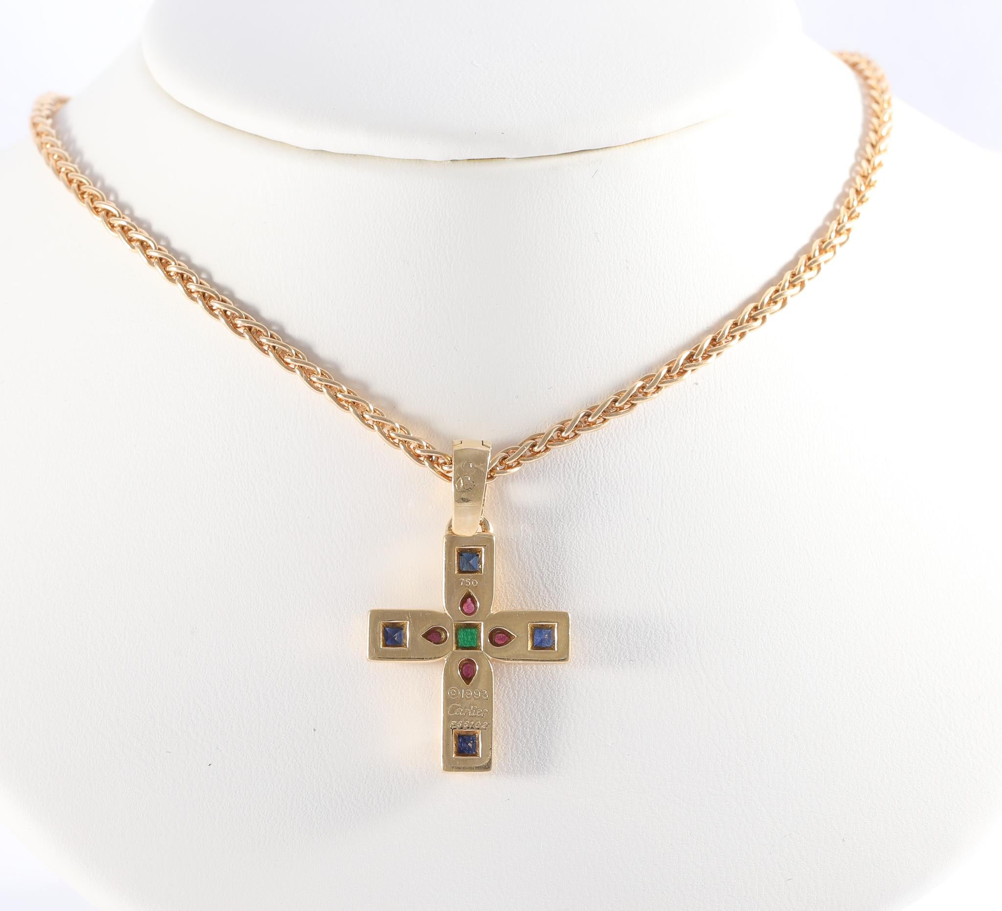 Cartier multi gemstone cross pendant featuring sapphires, rubies, and emerald, set in 18 karat yellow gold, suspended from an 18 karat yellow gold 17 inch rope chain. Both pendant and chain are stamped.