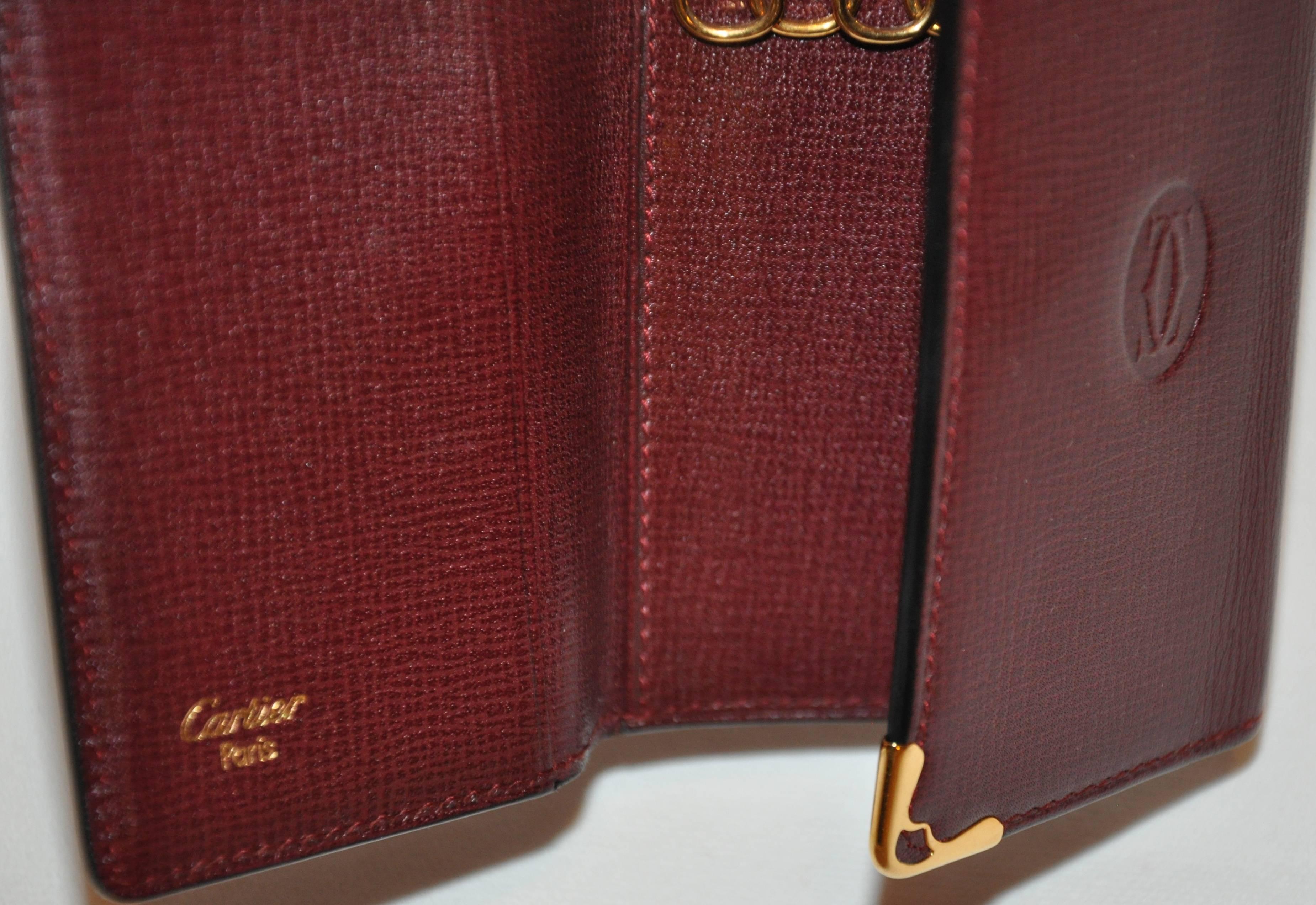        Der für Cartier typische Schlüsselanhänger aus bordeauxfarbenem, strukturiertem Kalbsleder ist an den Ecken mit goldfarbenen Beschlägen verziert. Das charakteristische Logo von Cartier ist elegant in die Mitte eingeprägt. Im geschlossenen