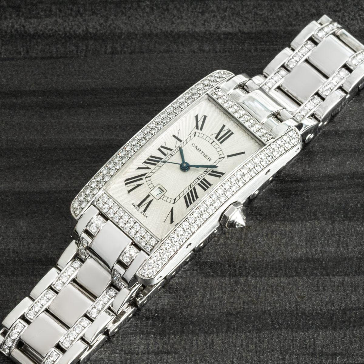 A striking white gold Tank Americaine by Cartier. Features a silver dial with Roman numerals, sword-shaped hands in blued steel, date display, and a Cartier signature hidden at 7 o'clock. Complementing the dial is a white gold bezel set with