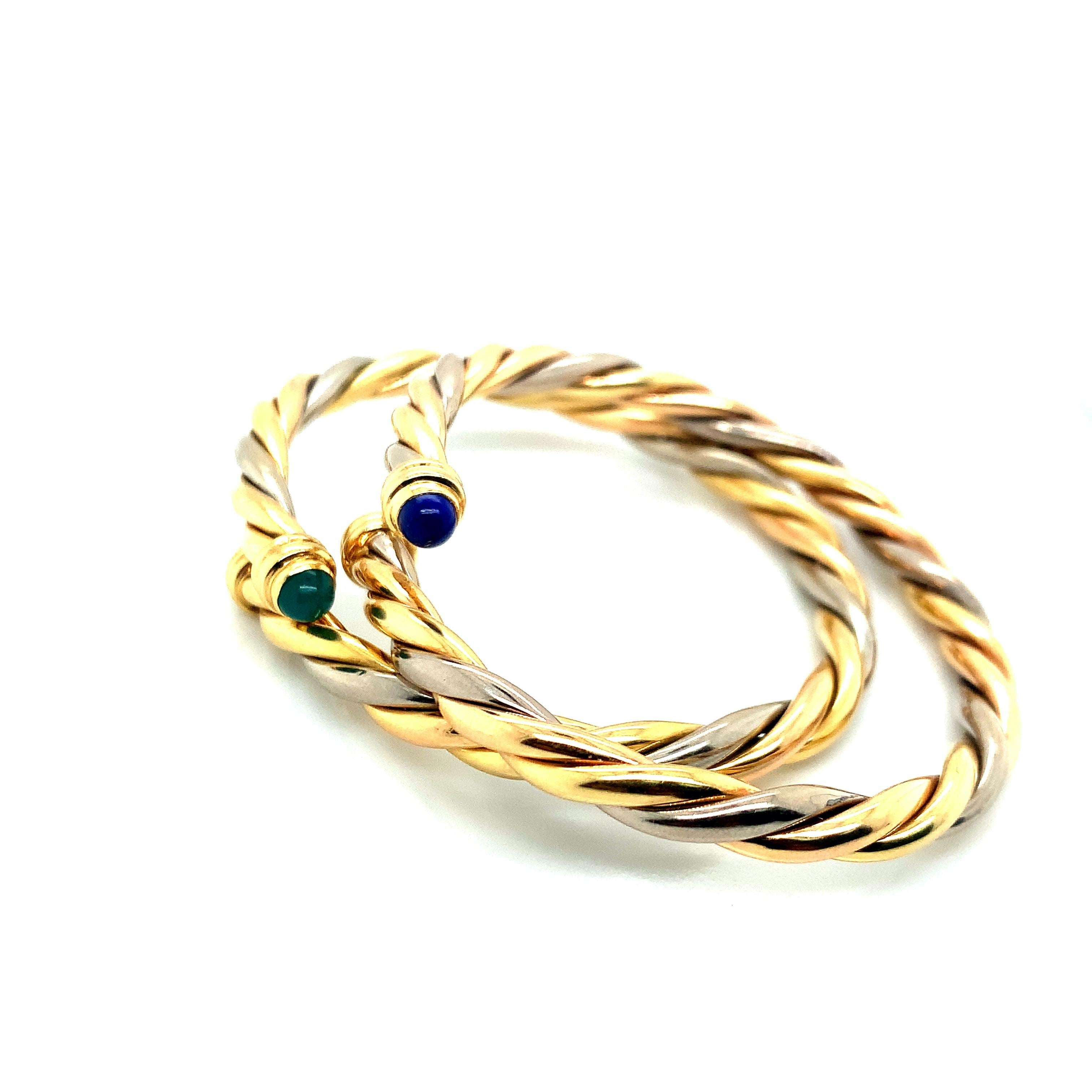 Two bangles that feature Cartier's classic design: the three tone 18 karat gold, including yellow, white, and rose. These two strands are twisted together to form a braid. Their ends are marked by either a sapphire or an emerald. Each bangle has its