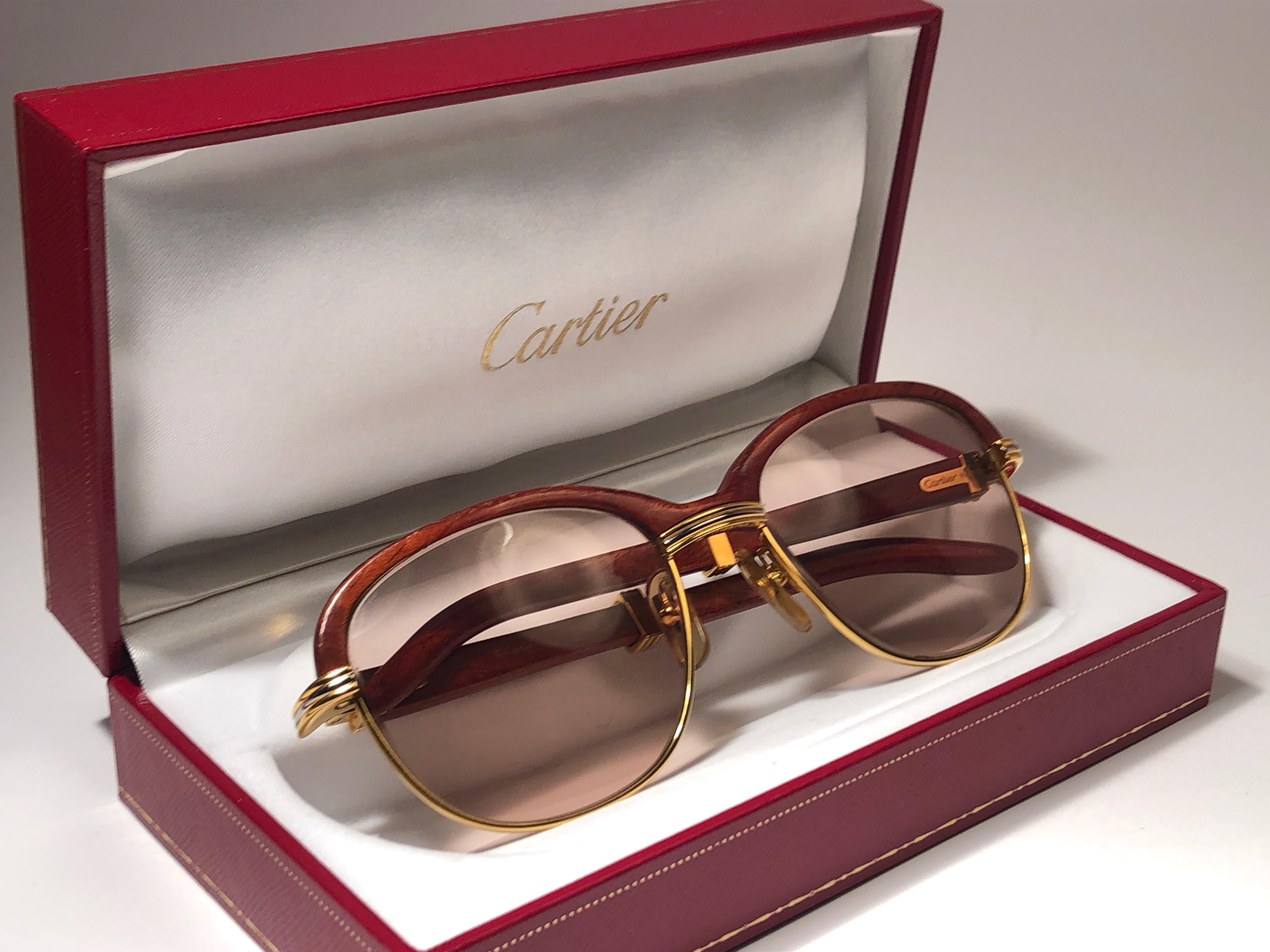 Lunettes de soleil Cartier Malmaison Palisander en bois précieux, originales et neuves de 1990, avec verres marron miel (protection contre les UV). 
Façade et flancs en or jaune et blanc arborant la prééminente façade en bois. Temples de palissandre