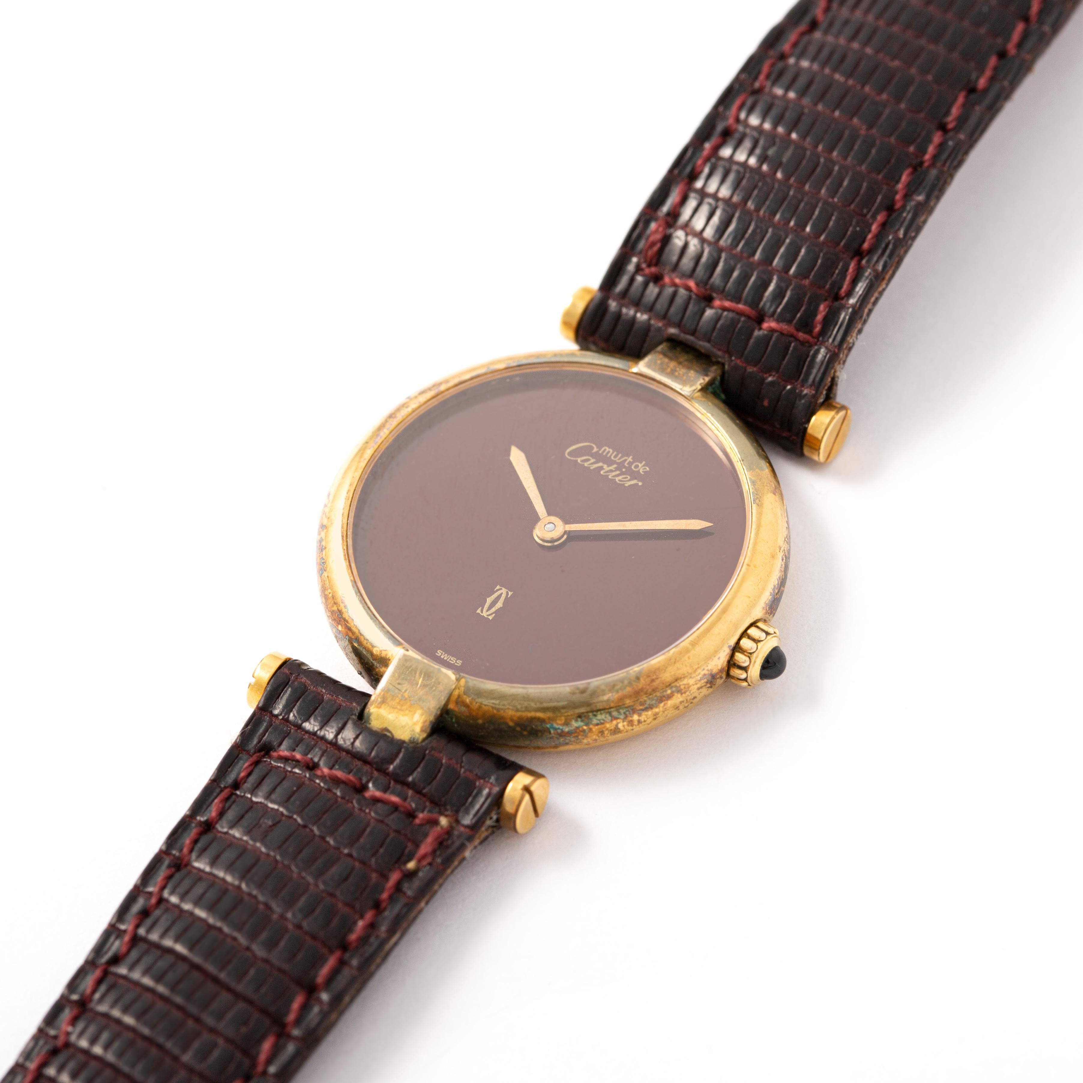 Cartier. Muss gesammelt werden. Runde Armbanduhr aus vergoldetem 925‰ Silber, burgunderfarbener Hintergrund.
Bordeauxfarbenes Lederband. Dornschließe. Saphir im Cabochon-Schliff auf dem Aufzugsmechanismus.
Quarzwerk. Signiert Cartier Paris.