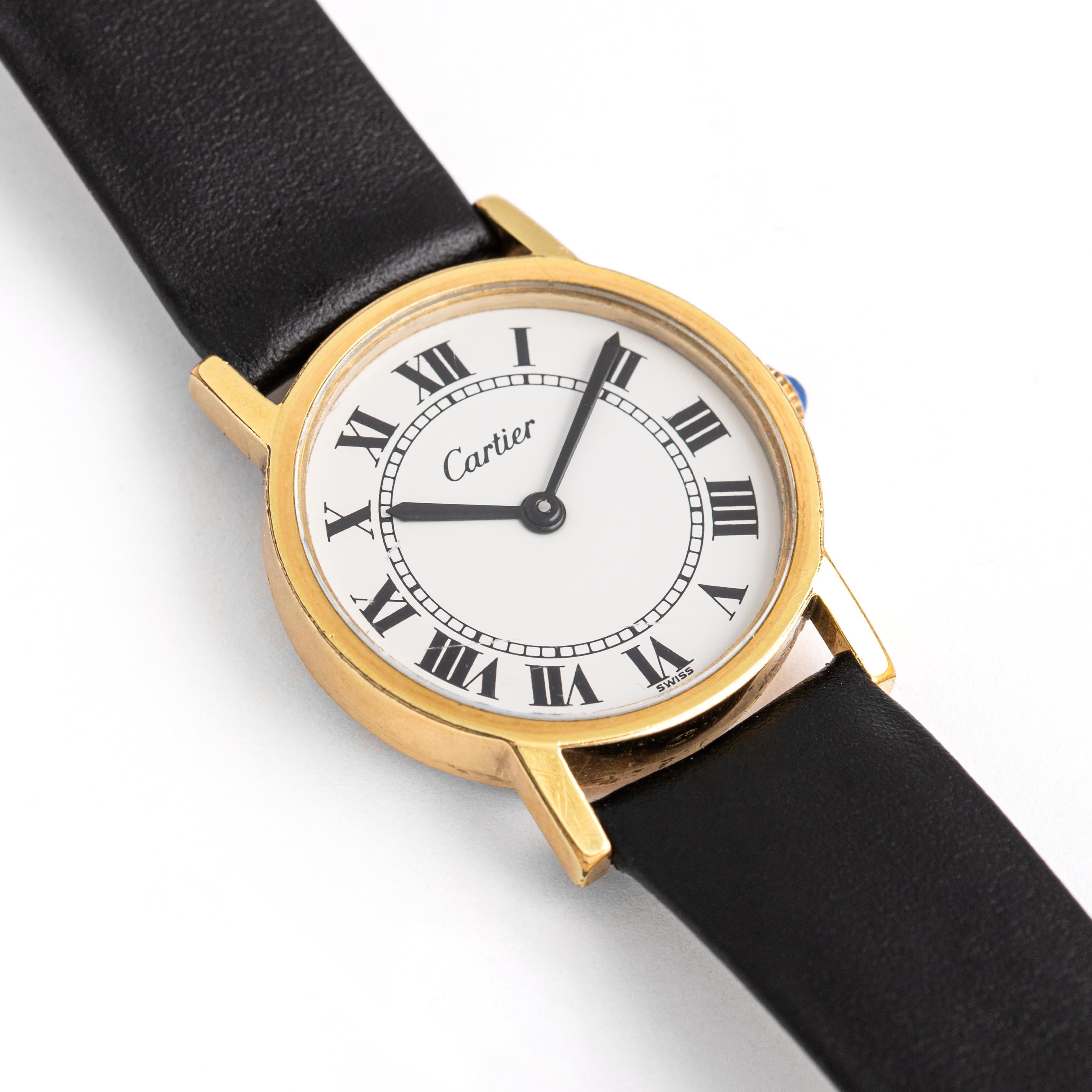 Cartier. Solo collection. Gold-plated metal wristwatch, round shape, white background and Roman numerals.
Black leather strap brand new and pin buckle. Cabochon-cut sapphire on the winding.
Quartz movement working. Signed Cartier. 
Numbered: