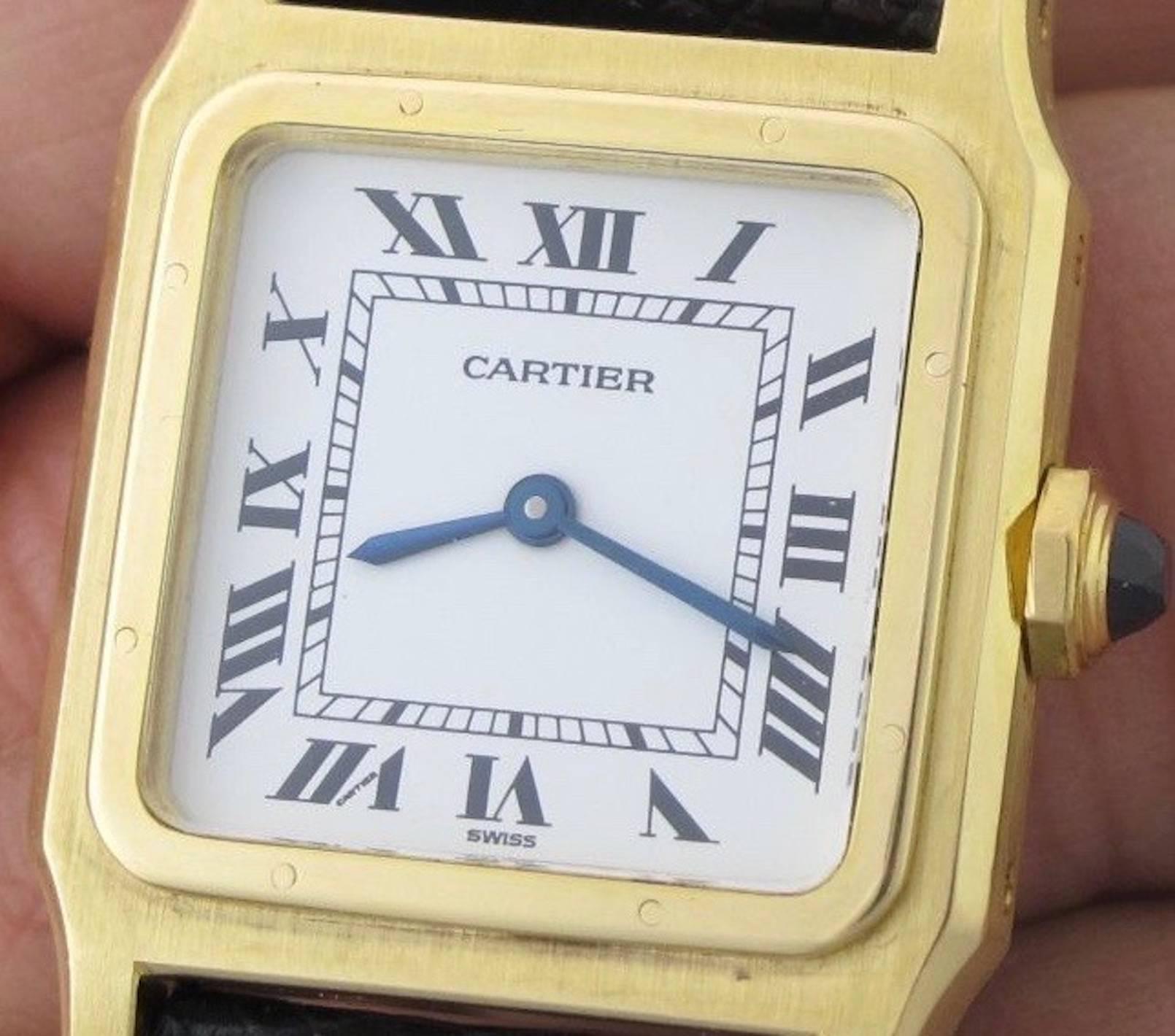 Cartier Santos Pre Owned Midsize Manual Winding wrist watch. White Dial with black Roman numerals. 18k Yellow Gold square style case with Blue Sapphire cabachon setting crown (27x36mm). Dark maroon strap with 18k Yellow Gold Cartier deployant clasp.
