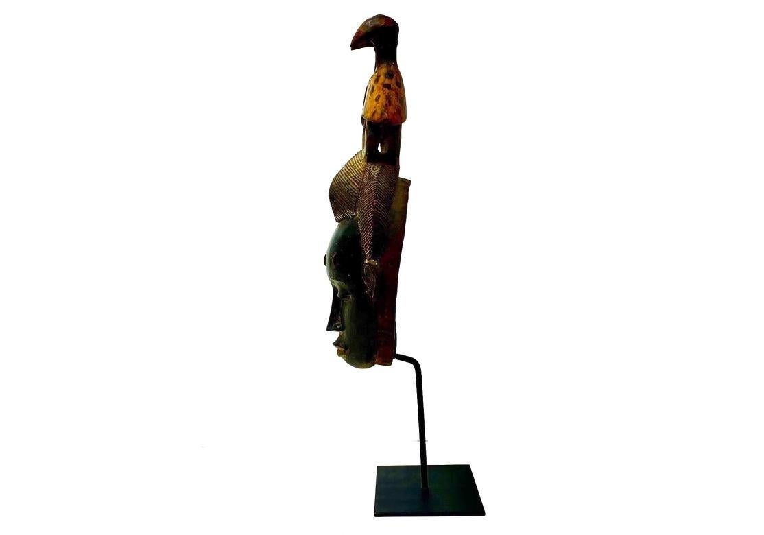 A strongly carved mask, likely Baule Tribe, on custom iron display stand. With fine serene features, scarification on the forehead, delicately carved hair and surmounted by two forward facing birds, likely protective. The interior displays numerous