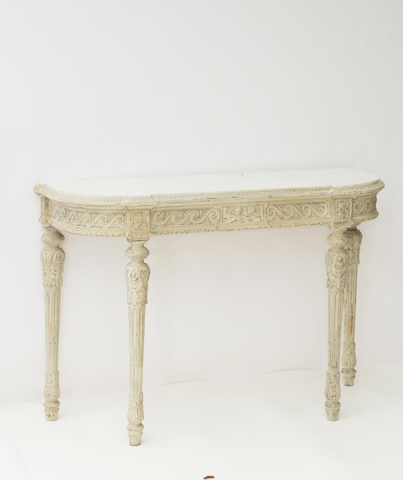 Console table, having a D-shaped top of Carrara marble, on a painted, conforming base, showing natural wear, with gadrooned border, its apron carved with evolute scrollwork, raised on round, tapering, stop-fluted legs, headed by foliate caps, ending