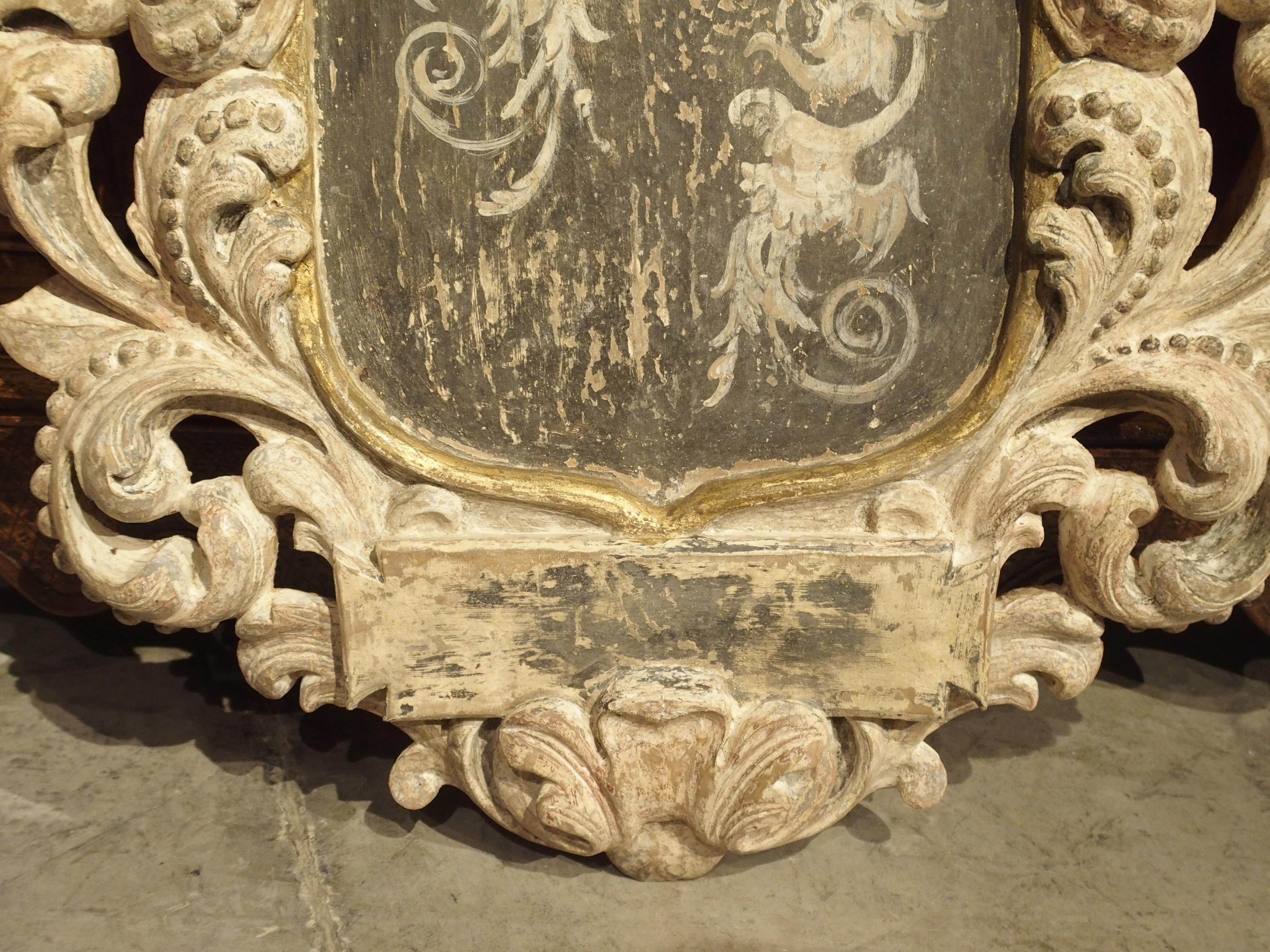 This wonderful hand carved and parcel paint plaque in the shape of a stylized cartouche is from Italy. The crown at the top is resting upon a shield form with painted demi figures of winged dragons. Surrounding the entire shield are thick scrolling,