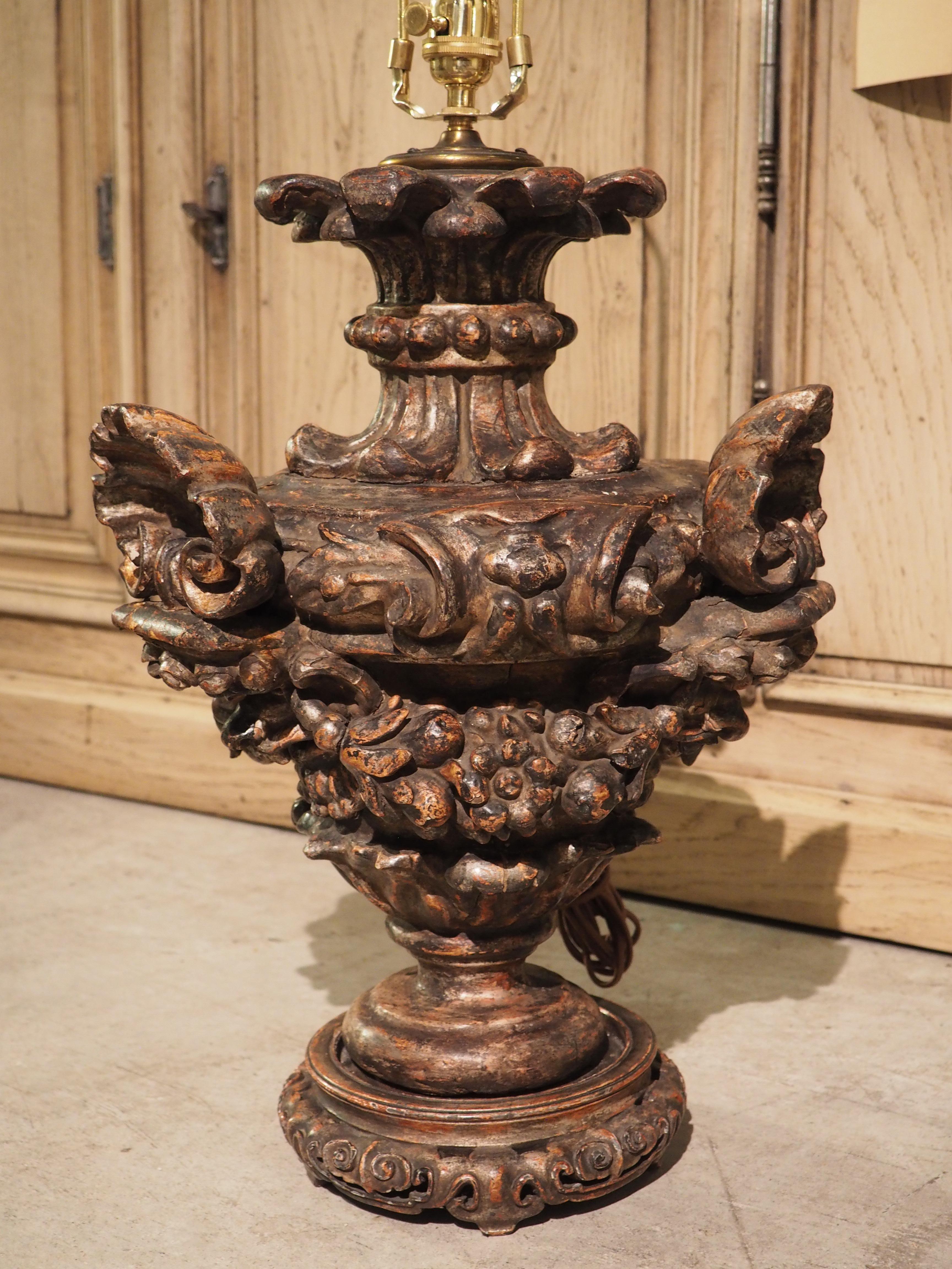 This highly carved polychrome urn from the 1700s has been repurposed into a unique table lamp with a cream shade adorned along the rim with banded S-scrolls in black. Three scalloped shells and three volute scrolls, in an alternating fashion,