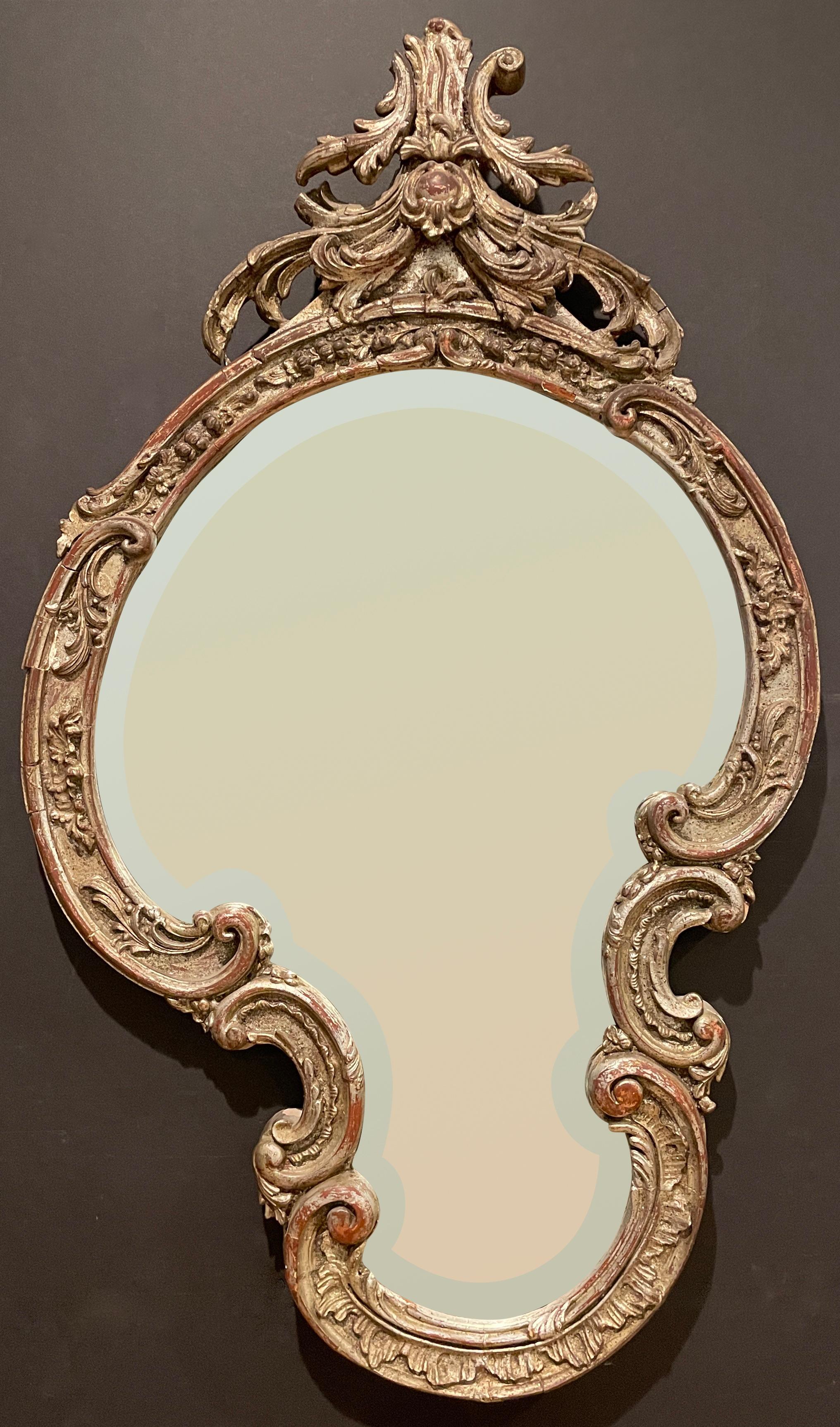 Beveled mirror in the Rococo style with hand carved elements throughout. Free-form shape with worn silver gilt finish and original antique mirror.