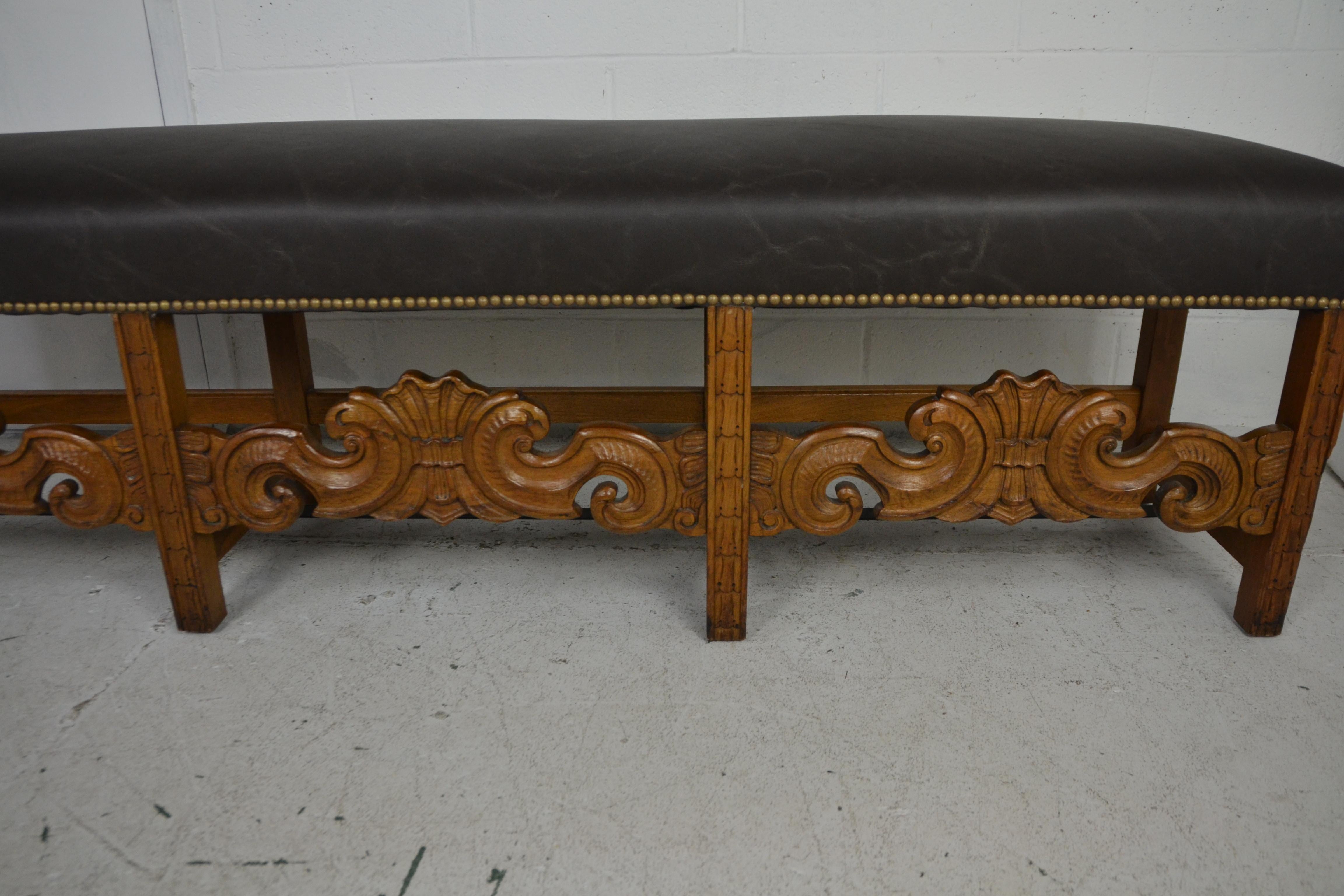 An extra long carved bench in the Louis XIV style. Newly upholstered in brown leather. Brass studs.