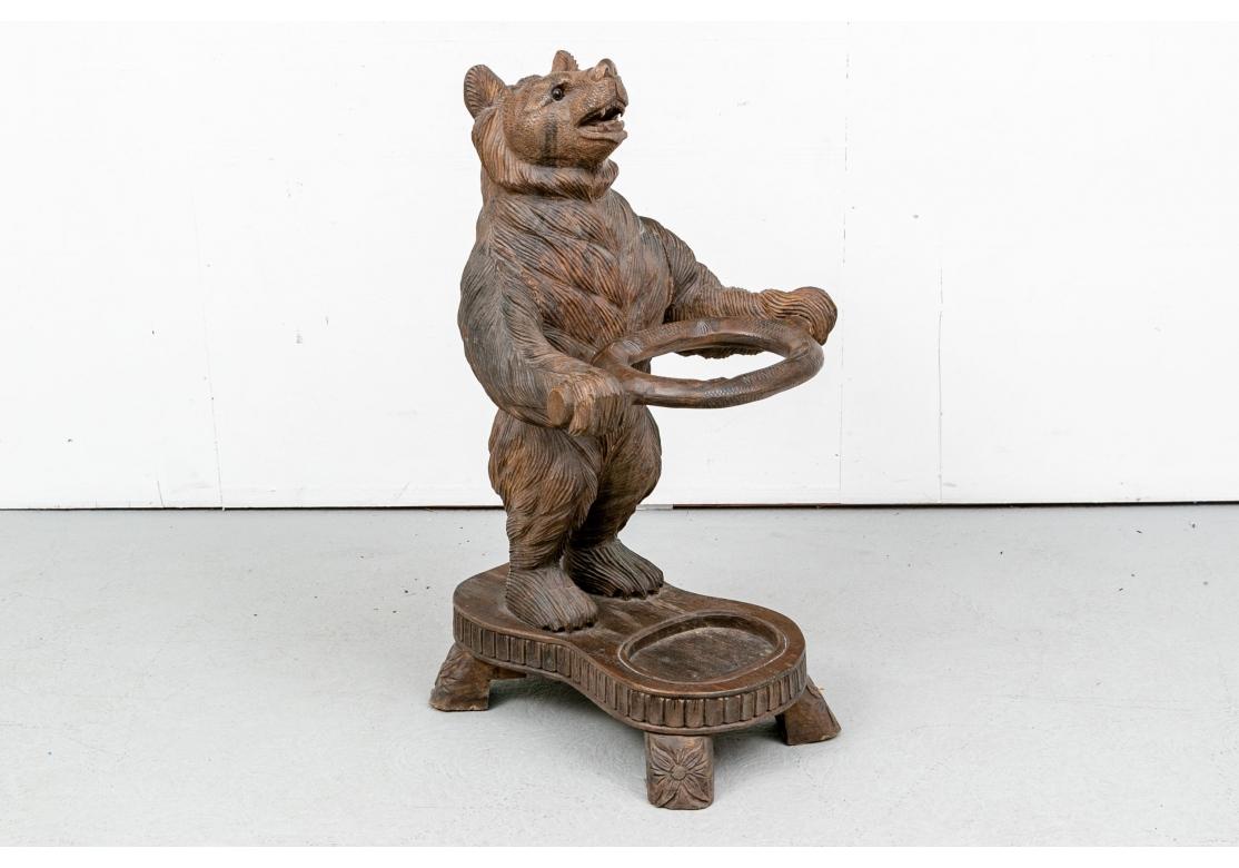 A very solid black forest style bear carving serving as a cane and umbrella stand. A standing bear figure holding the open oval support for umbrellas and canes. The shaped base with an oval depression in the front, raised on blocky feet with carved