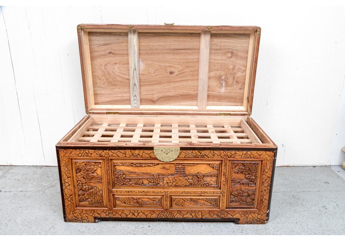 Tronc en bois de camphrier avec couvercle à charnière, lourdement et minutieusement sculpté de panneaux en relief représentant des paysages, des arbres, des pagodes, des motifs feuillus sur fond de fleurs. Le coffre s'ouvre à l'aide de poignées