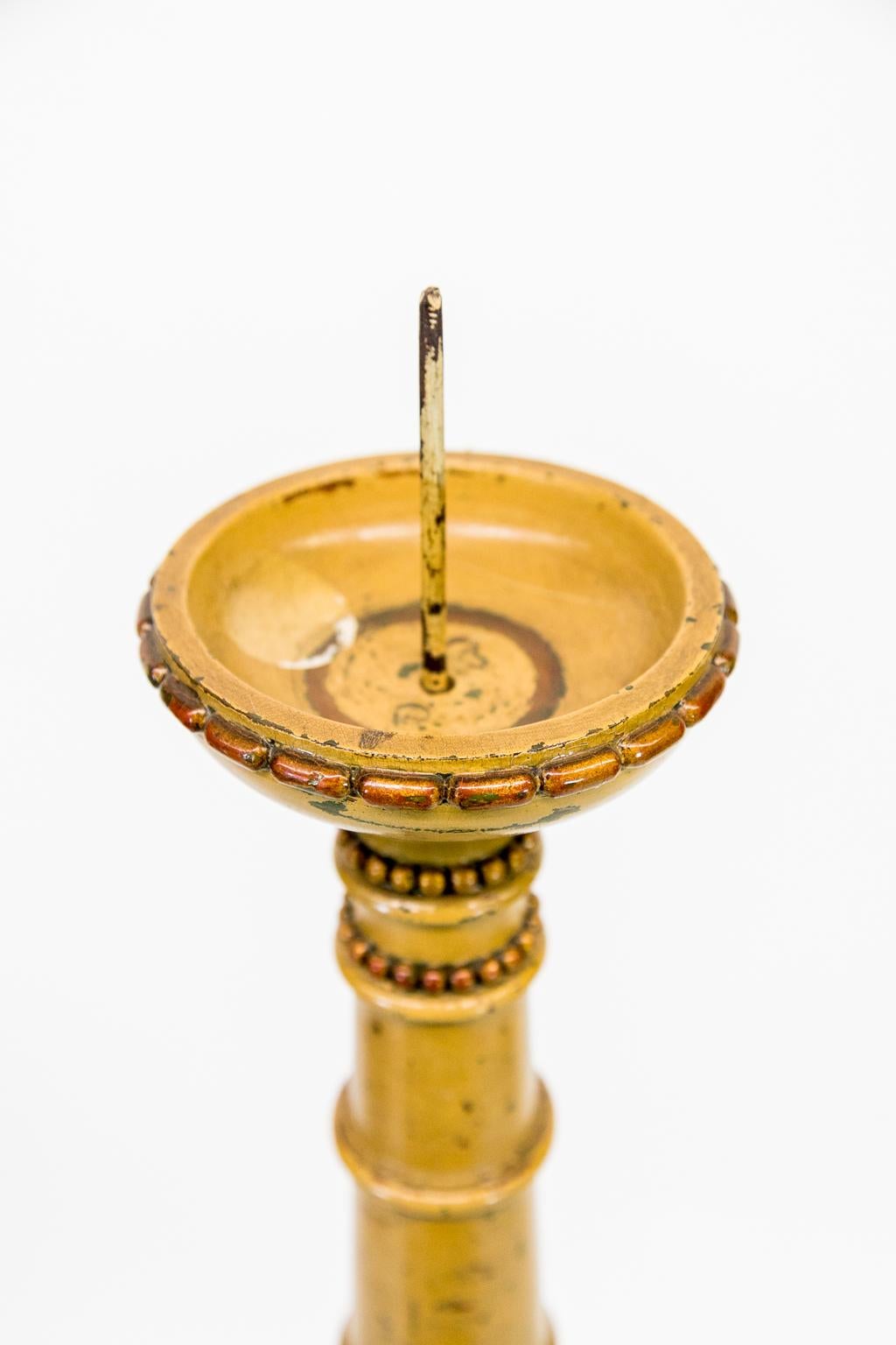 This candlestand has a four-inch steel pricket. The top edge has a repeating capsule border. The upper stem has turned rings and repeating bead carvings that surround the stem. The center of the stem has acanthus leaves carved in high relief. It has