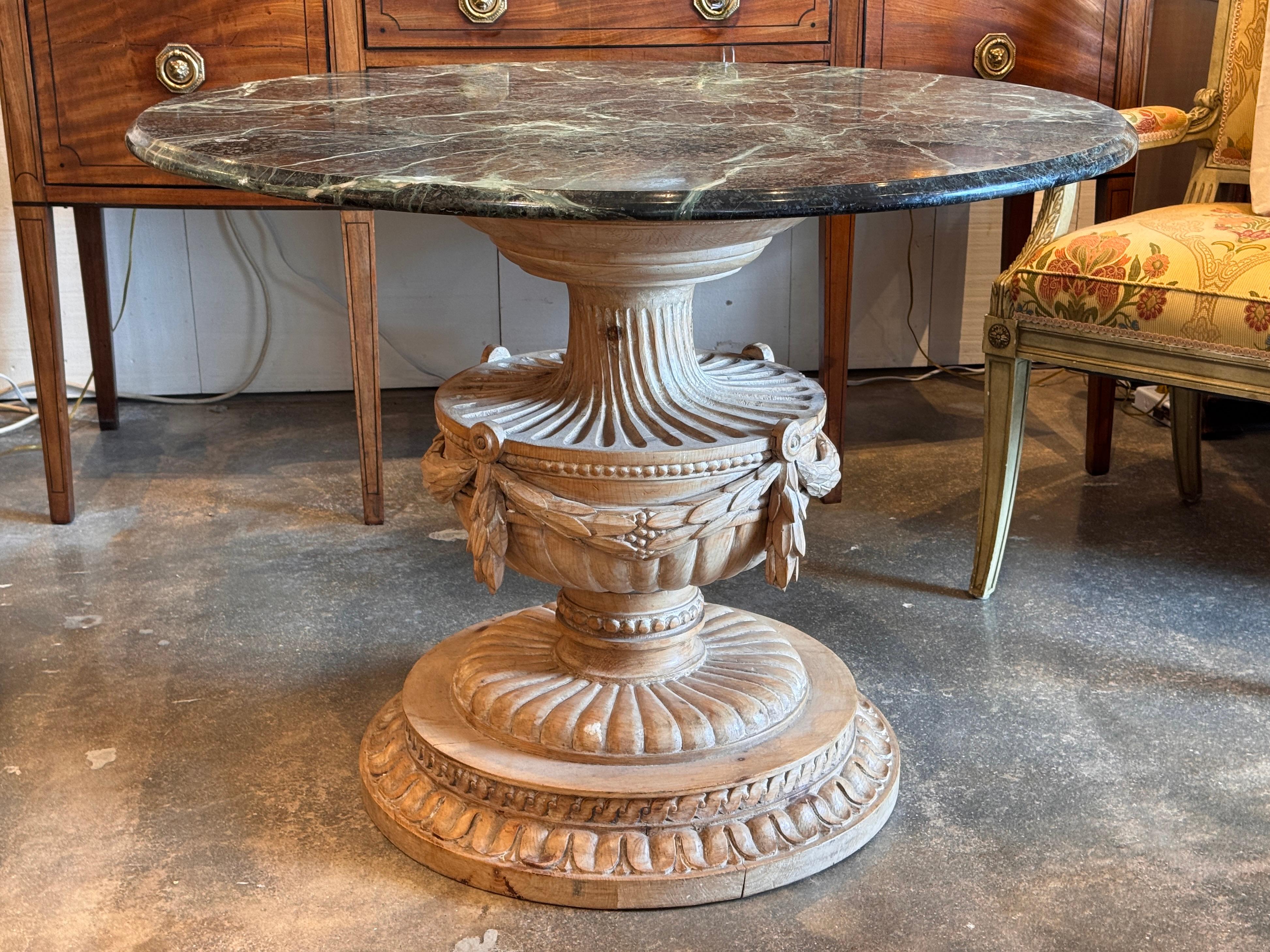 Carved center table featuring a round deep green with white veining marble top supported by a hand-carved wood pedestal with leaf and ribbon motifs over a stepped circular base. The close-up highlights the crisp carving and natural wood finish