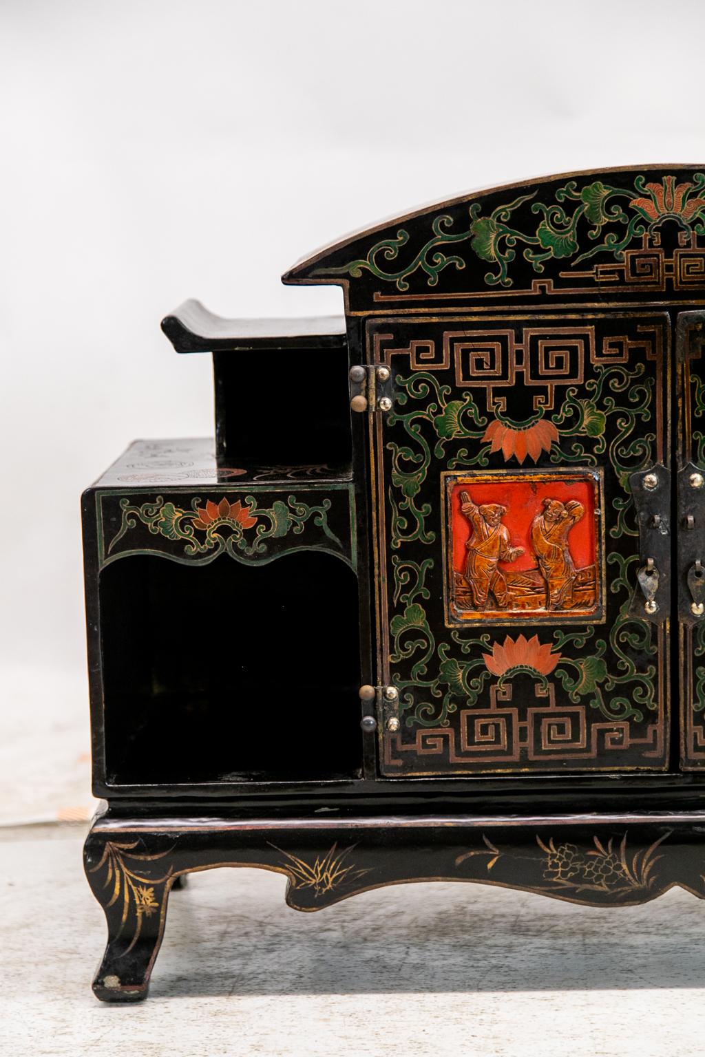 This carved Chinese cabinet is painted with red, green, and gold geometric and Asian arabesque motifs. The center panels have carved Chinese figures. The apron and legs are painted with gold leaf and floral decoration.