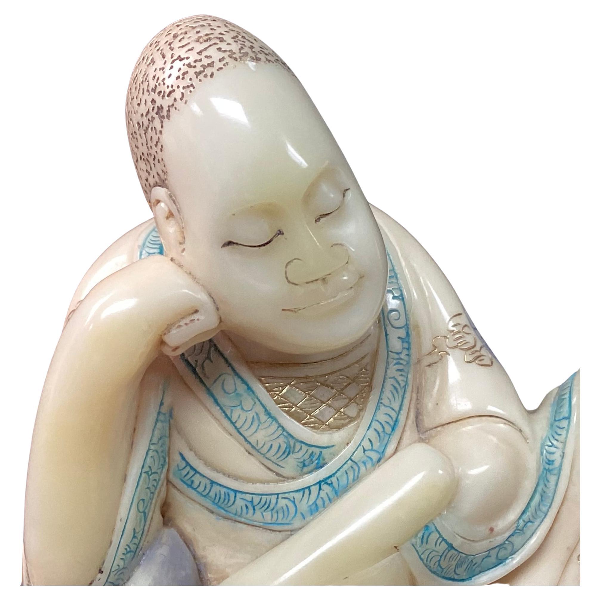 19th century carved Chinese Soapstone Arhat figure in the style of the Qing dynasty. Intricately carved figure is in seated position, hand to face, wearing a robe with aqua and dark blue symbols on cream background.