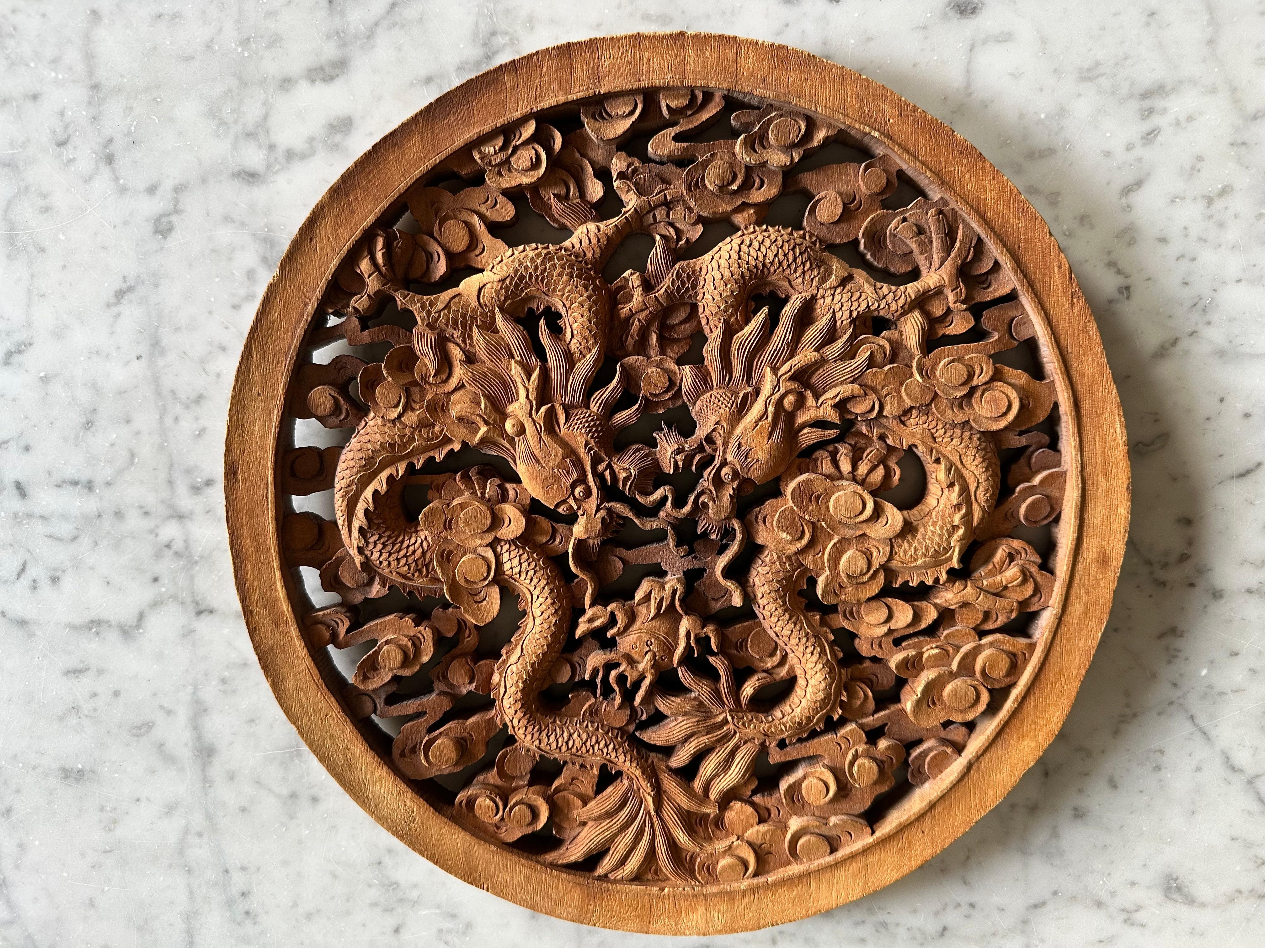 This round carved panel from China, made of rubber wood, features a traditional double dragon motif in openwork relief. The two dragons are depicted in motion, their sinuous bodies intertwined in a balanced and symmetrical composition. The carving