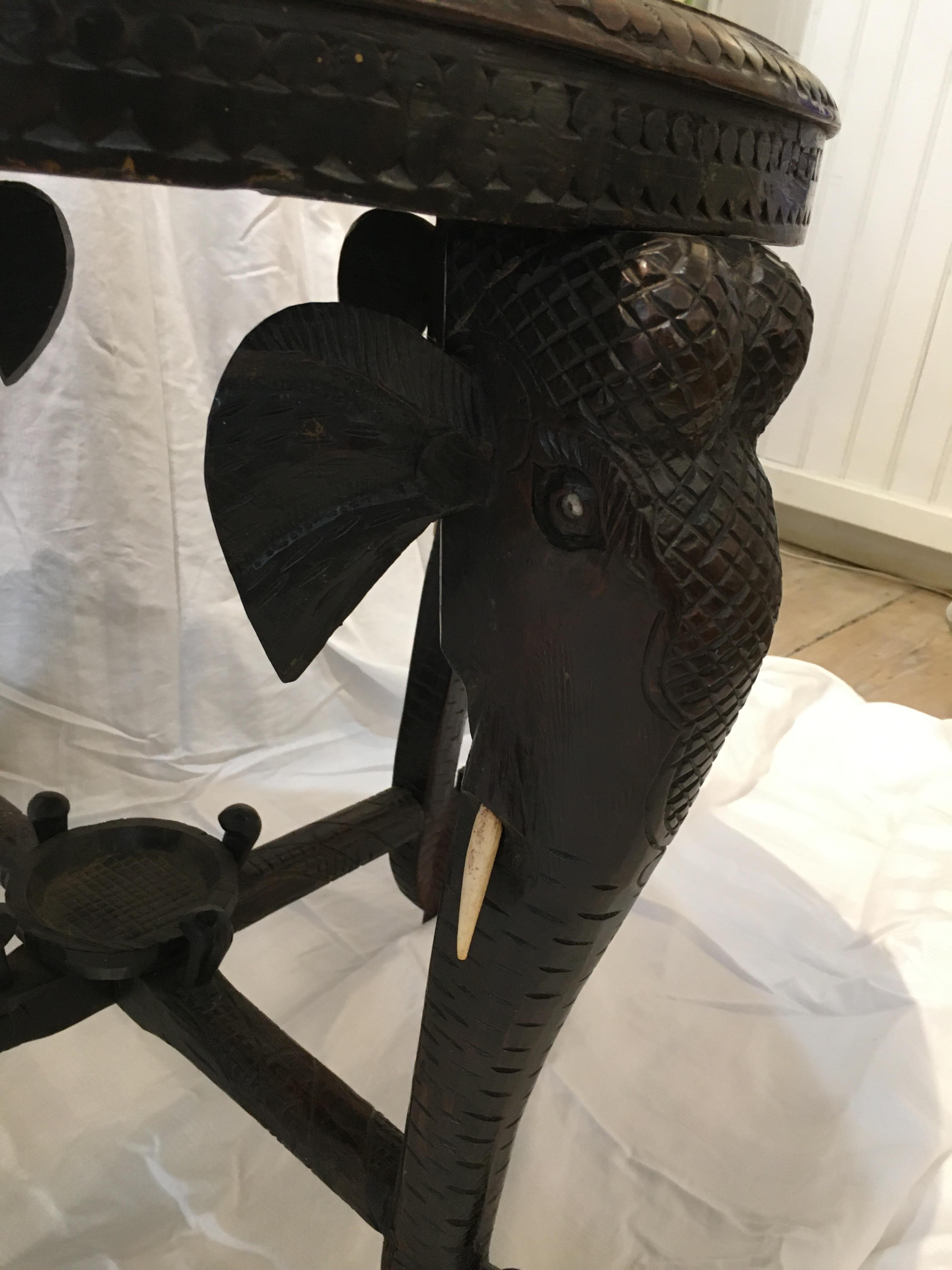 An intricately carved ebonized wood side or center table with bone inlay and featuring elephant head, tusk and trunk legs. Carved leaf patterns around the top border and a center full body elephant. The inlays are flowers and vines along with two