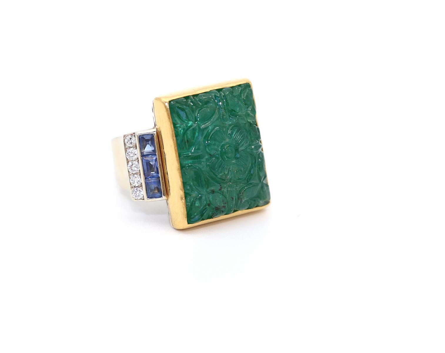 Carved Emerald Sapphires Diamonds Ring.
Massive and very rare precious Emerald artistically carved with floral ornamentation. Framed in 18K Yellow Gold. Underlined by two rows of Sapphires and fine Diamonds on each side. The sheer size of the ring