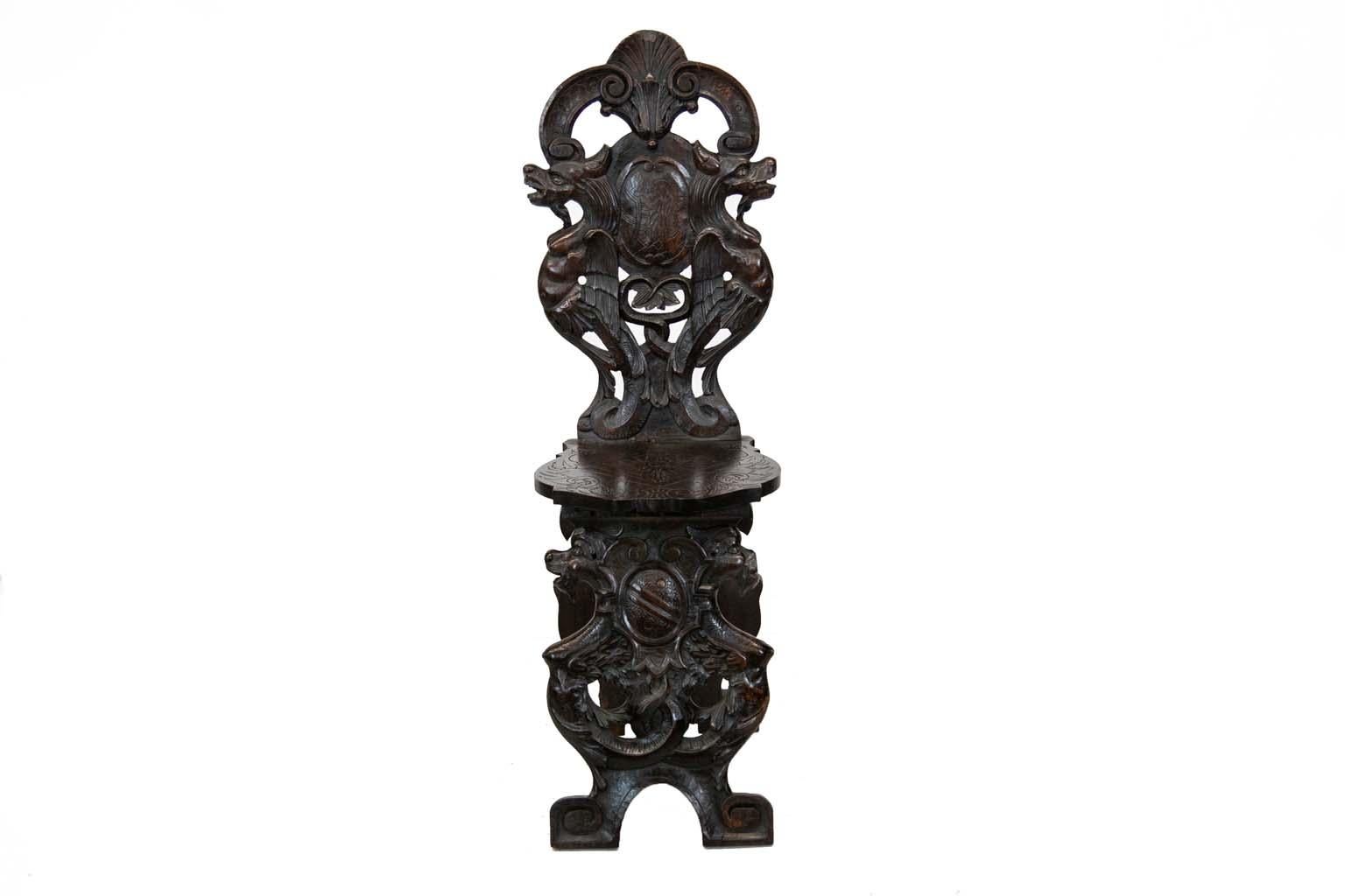 Carved English oak hall chair has a plank seat carved with stylized floral arabesques on the edge. A center panel is carved with framed stylized leaves. The center cartouche has a carved lion which is framed by winged gargoyles. The seat support