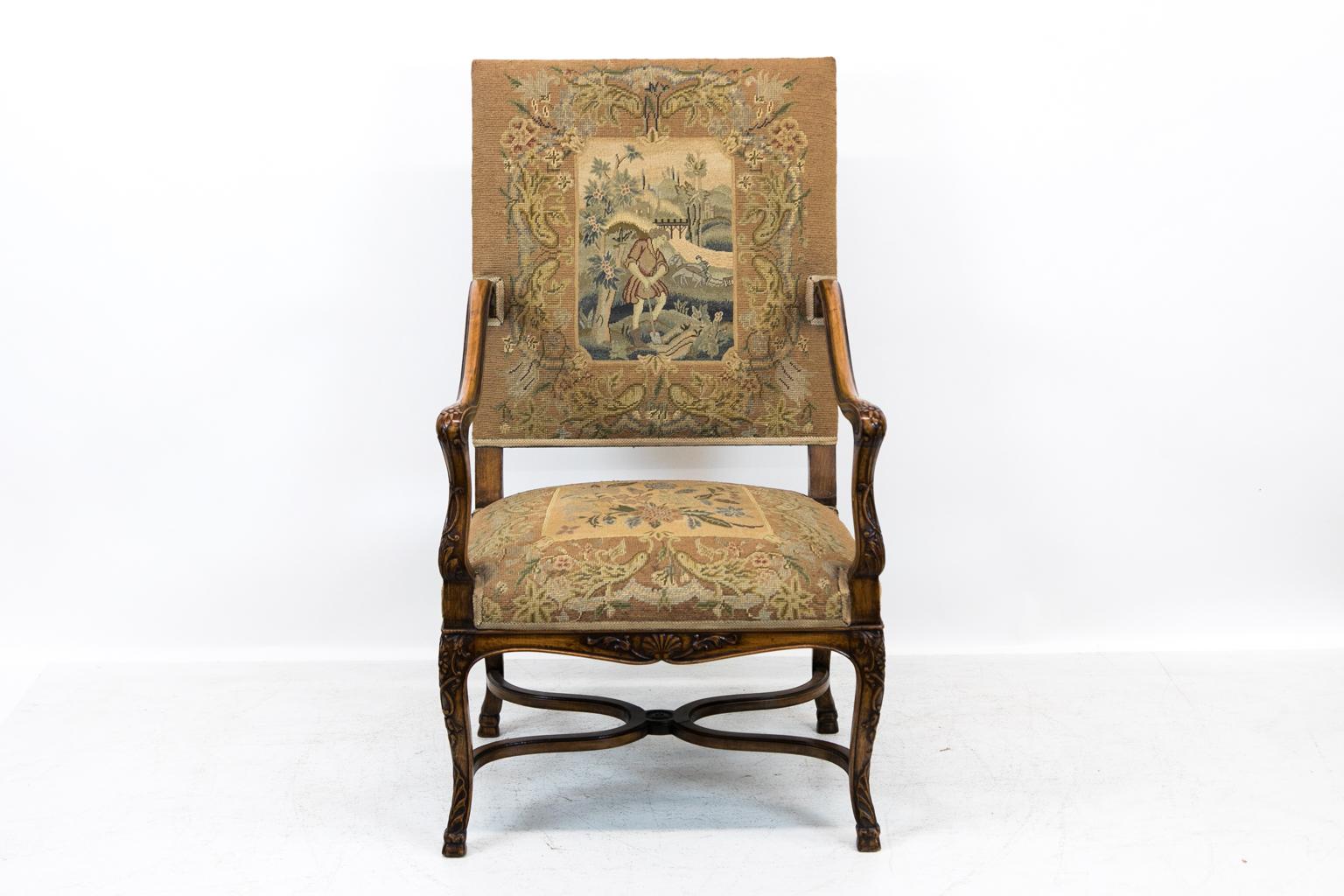 Carved French armchair, has shaped scrolled arms with acanthus carved supports and legs. The center panel is stitched petit point pastoral scenes and flowers. The base has a molded cross stretcher with a floral medallion, and the apron is carved