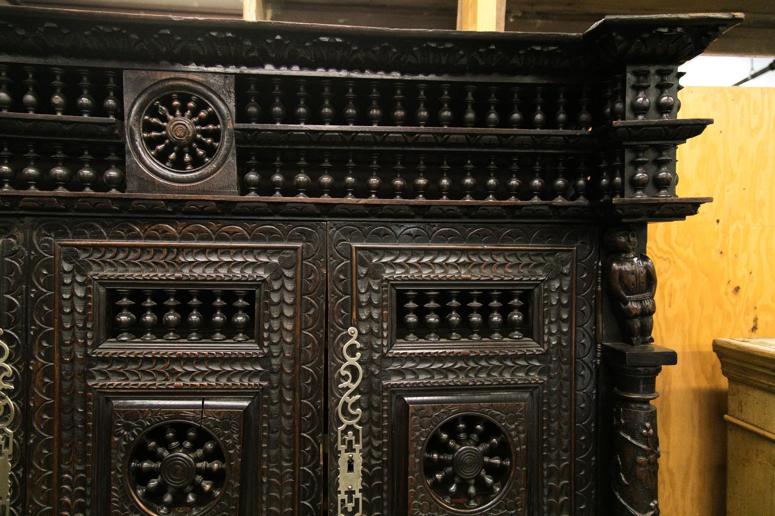 This French buffet deux corps is profusely carved with repeating stylized leaf designs with free standing carved grapevine and grape motif columns in the upper section that support carved figures of men. The upper cornice has a double open