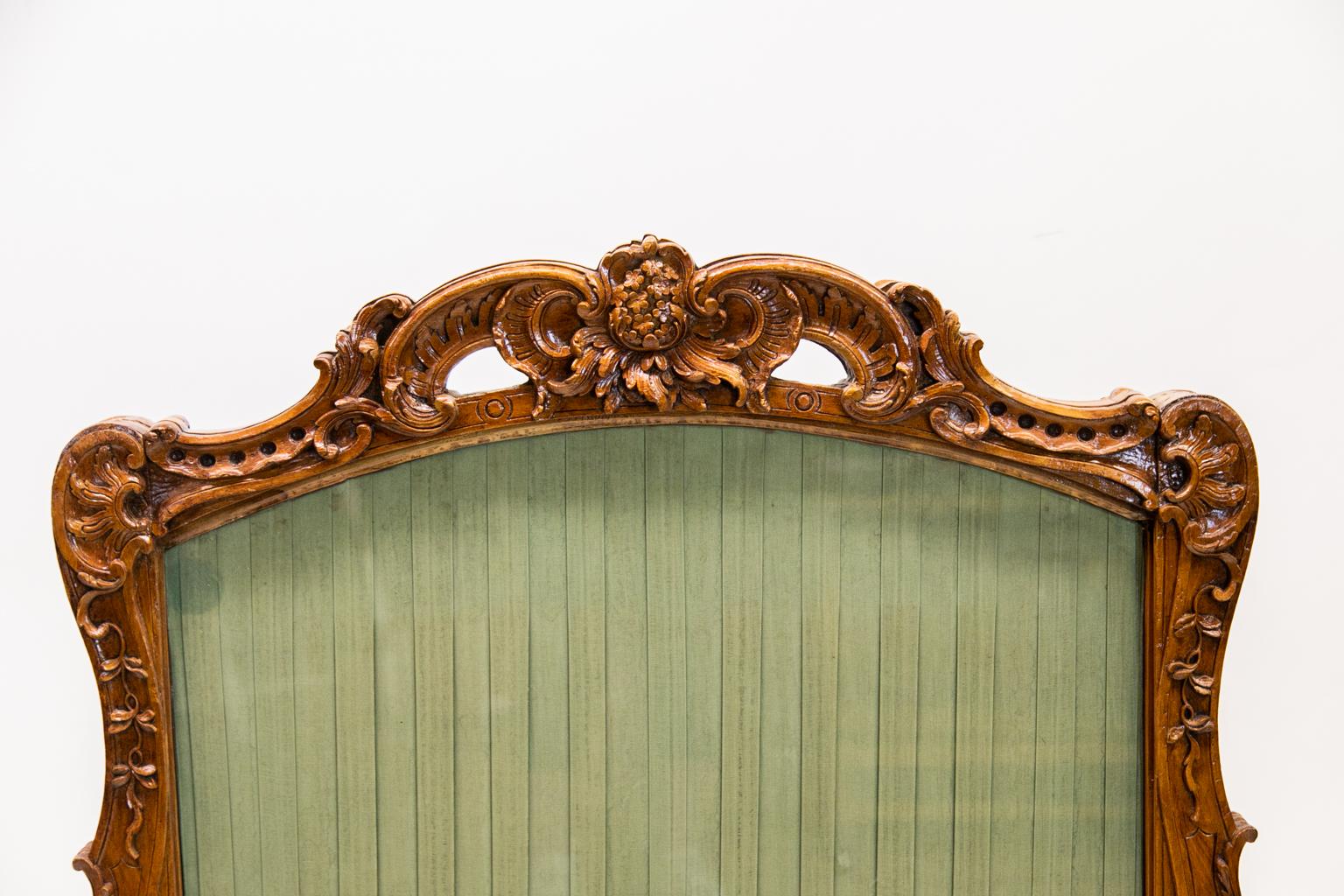 This carved French fire screen is carved on the front and back with identical carvings of floral and arabesques designs. The front and back legs terminate in scrolled feet. The central panel as a pleated green fabric with protective glass on the