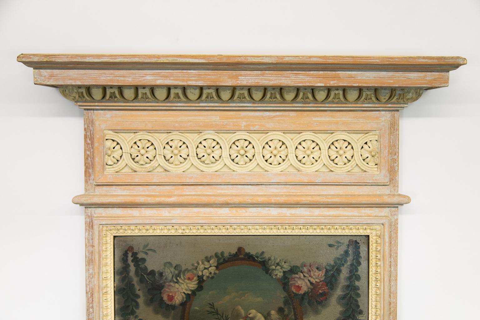 This mirror is painted with a cream color paint that has been intentionally distressed. The cornice has egg and dart molding carved in high relief. The frieze has a carved interlaced floral motif framed by rectangular molding. The painting above the