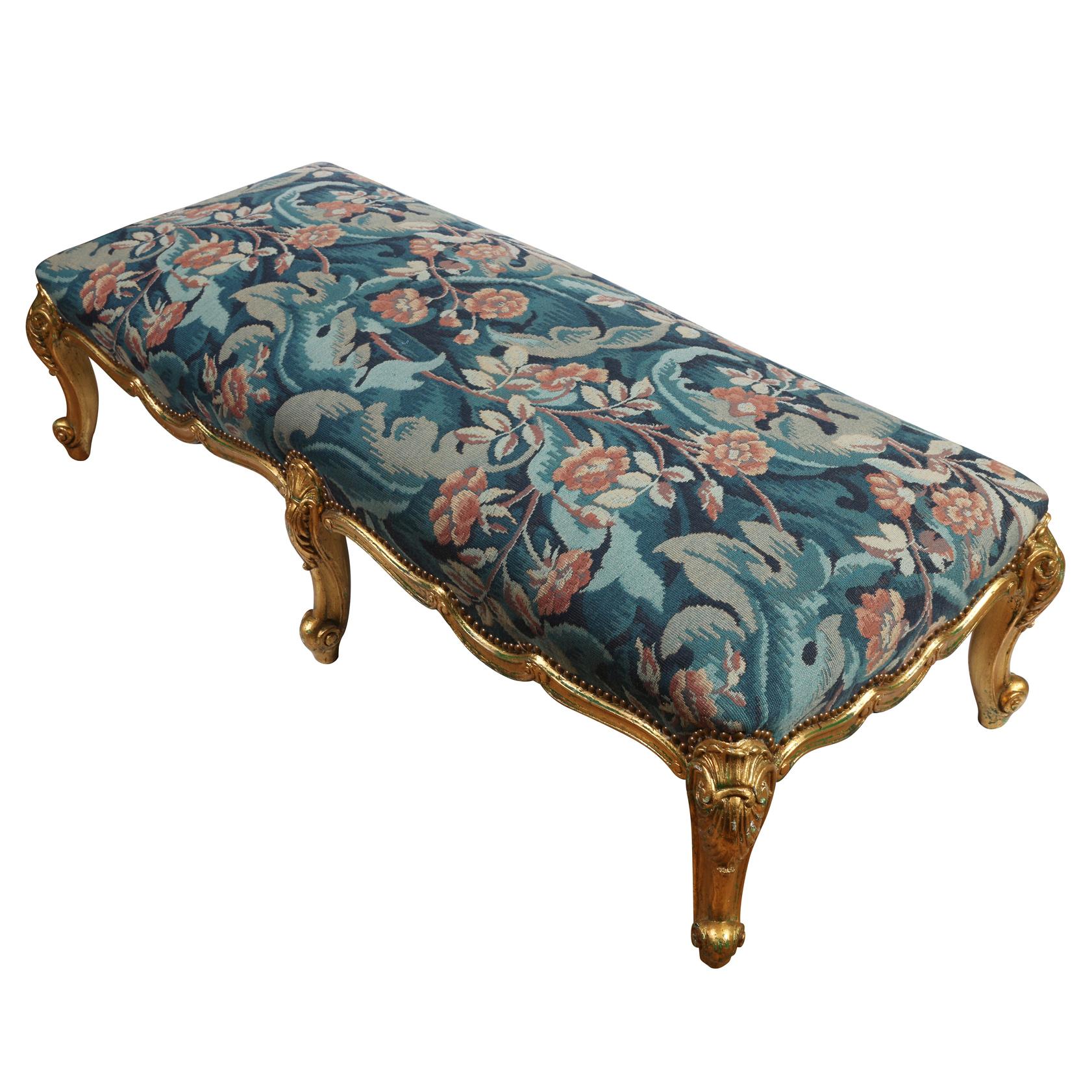 A Louis XV style oversized giltwood carved bench with six legs and upholstered in blue tapestry fabric with leaves and coral flowers.
