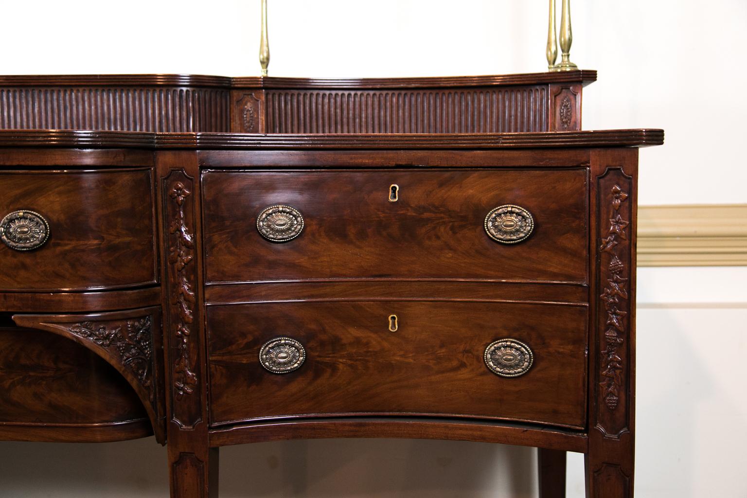 Carved Hepplewhite sideboard, with a sliding tambour plateau and a brass gallery. The front legs and center drawer are carved with oak leaves, acorns, and grapevines. The fronts are made of flame grained mahogany.
  