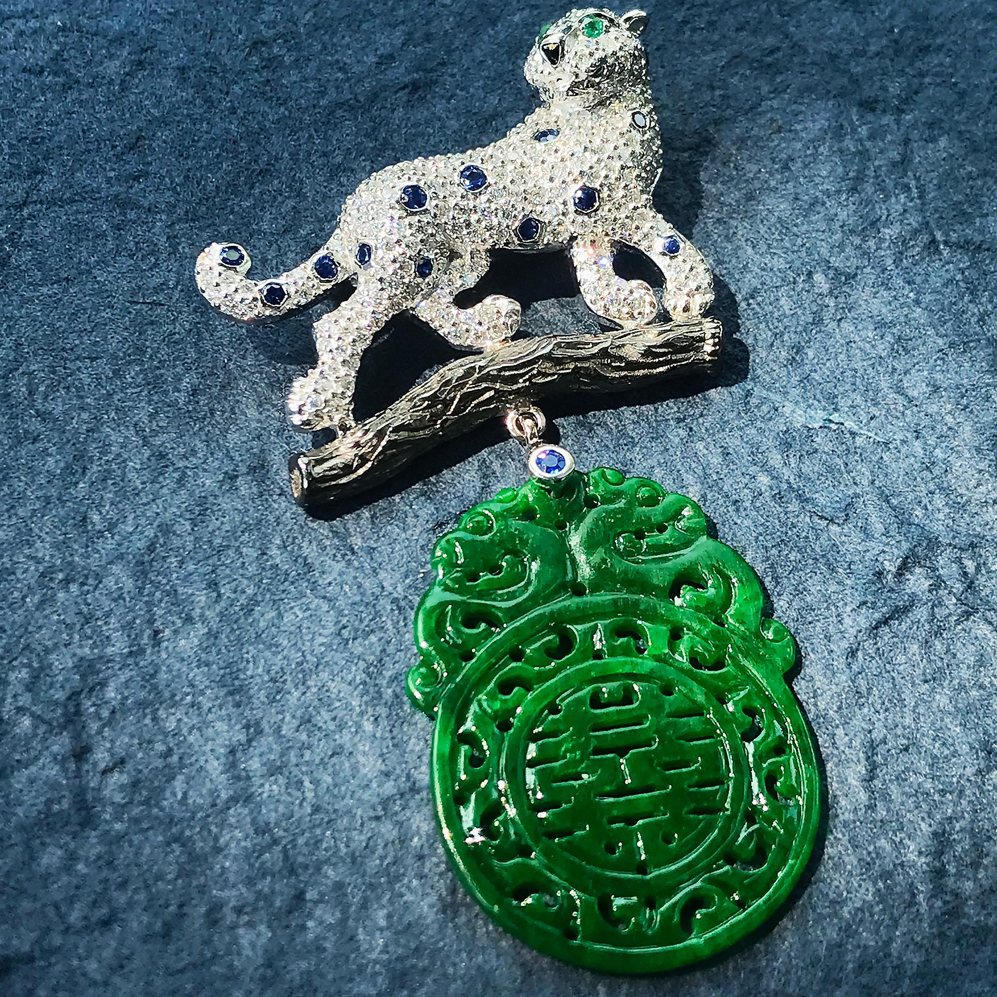 Diamond panther climbing on a branch brooch crafted in 14k white gold, its body set with round diamonds accented with sapphire, partially rotating head, with eyes set with emeralds and onyx nose. Below is a 11.9 carats beautifully carved jade. A