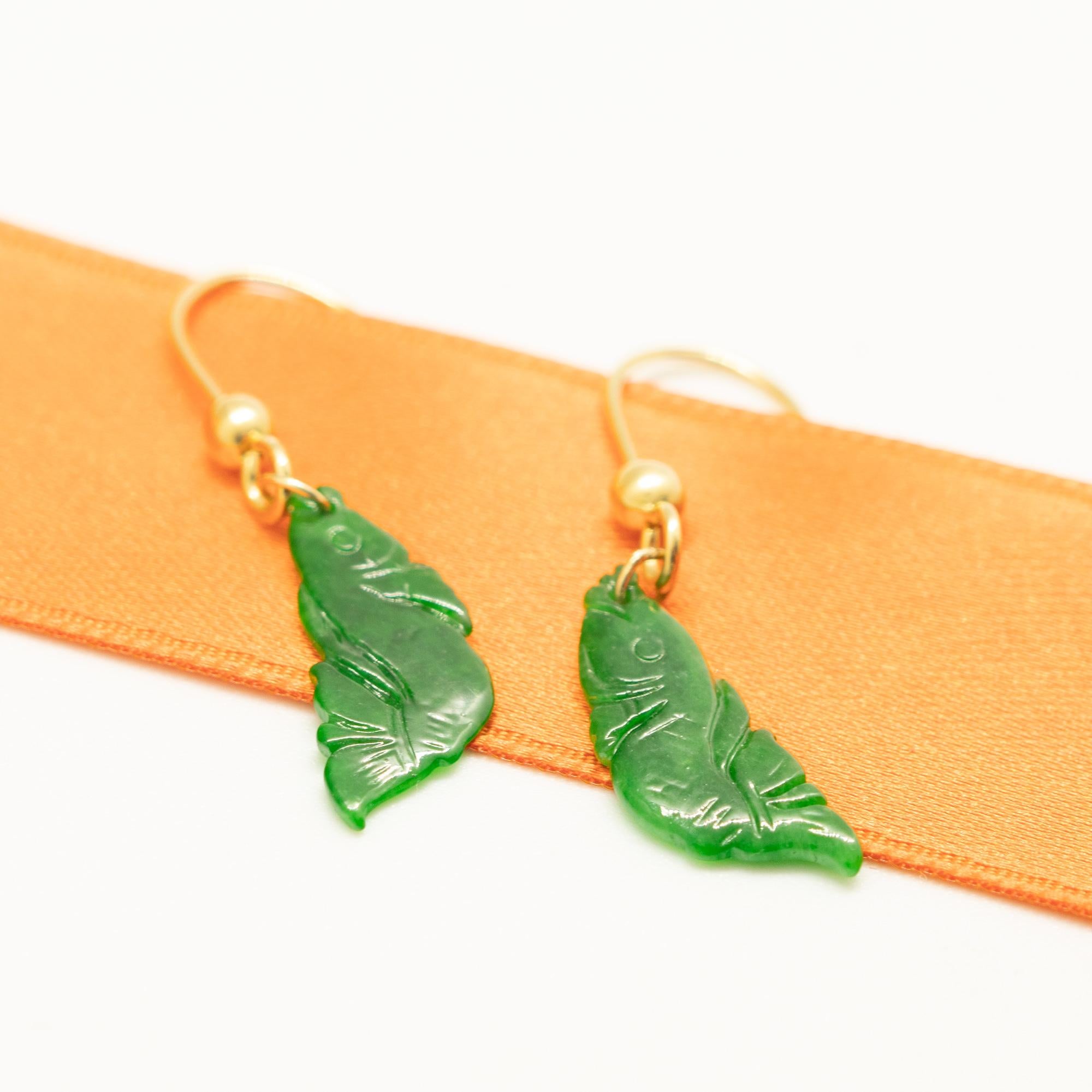 A stunning 9 karat yellow gold earrings ending in a unique natural carved jade stone. The green precious stone is carved in a fish shape with a flower tale. Long, dangle drop earrings created for a bold and delicate look. 
 
These earrings are
