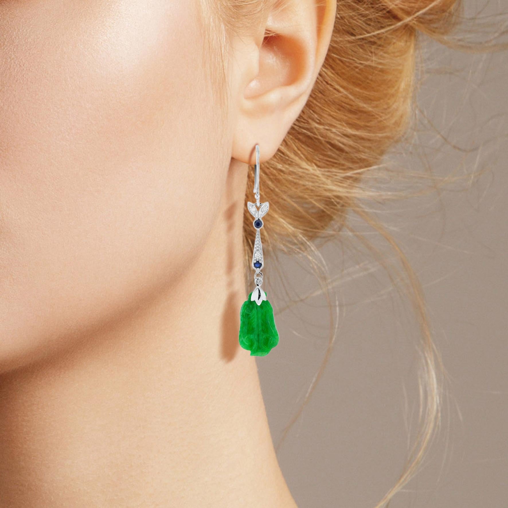 Die natürliche Jade in diesen schönen Ohrringen hat eine ganz besondere Form. Beide Stücke haben eine kräftige grüne Farbe und gute Schnitzereien. Die Jade hängt in einer Fassung aus 9 Karat Weißgold mit einem Diamantblatt und runden blauen Saphiren