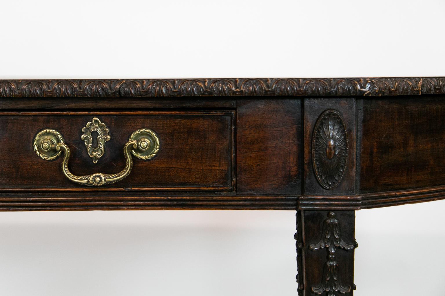 This carved mahogany console table has a straight front with rounded ends. The top edge has applied carved molding with a stylized leaf motif. The top of the legs have oval medallions with applied bellflowers below. The apron has reeded molding that