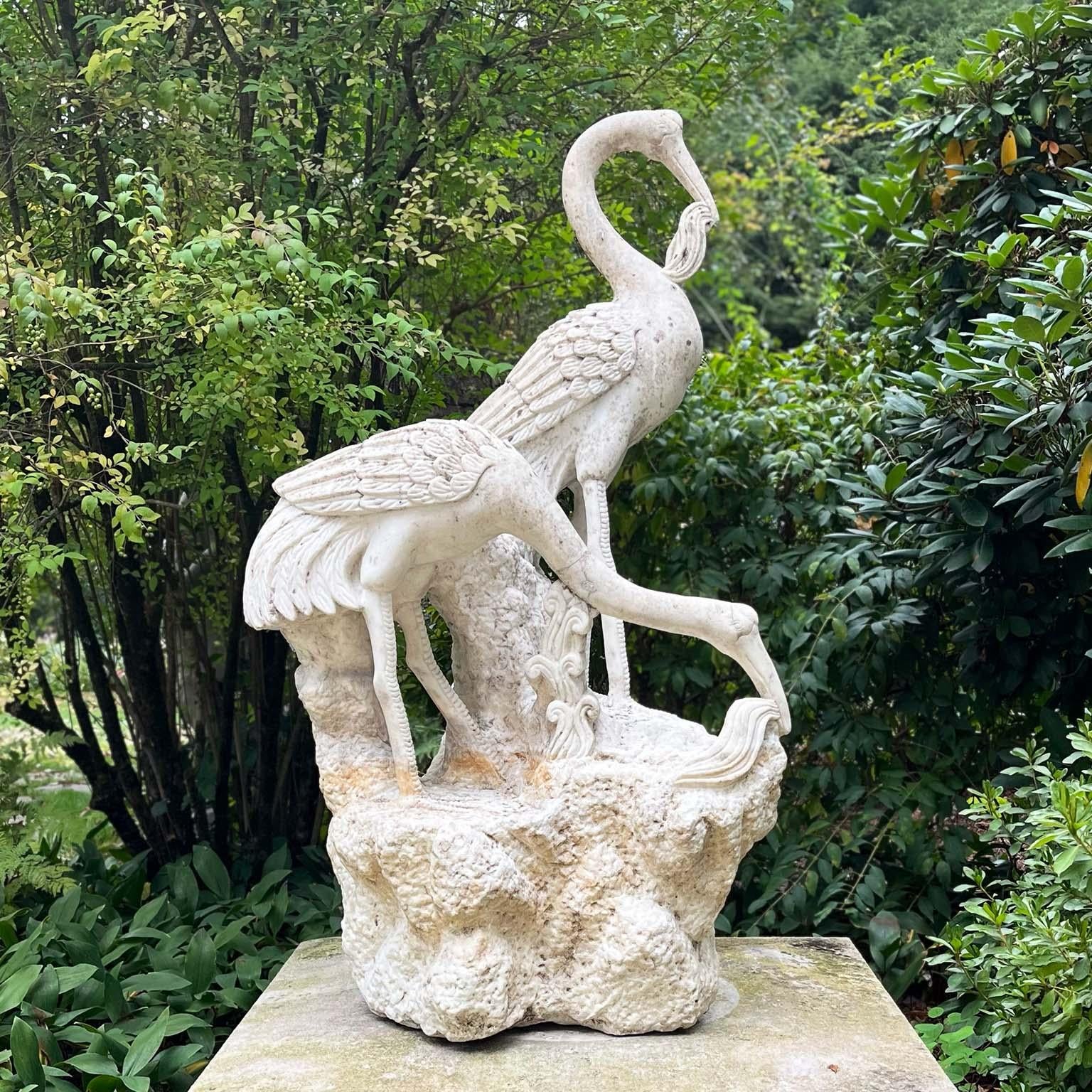 Groupe en marbre sculpté représentant deux grues sur une base rocheuse, l'une des grues, tête baissée, récoltant des plantes aquatiques, l'autre, debout, tenant des plantes aquatiques dans son bec, les deux oiseaux ayant des plumes finement rendues.