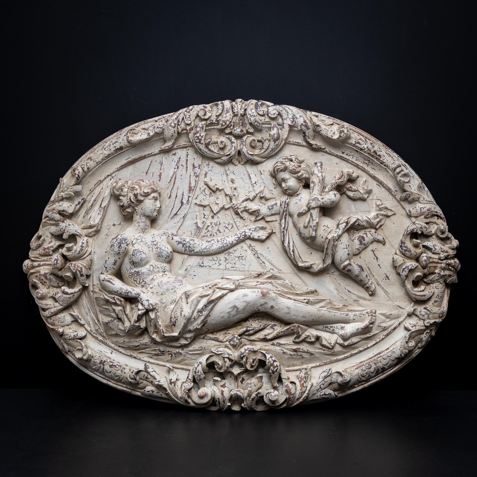 Carved relief in medallion form with rocaille decoration on the frame and depiction of Venus reclining, holding out her hand to Cupid armed with lightning bolts. The relief has been painted white and given an antique patina.
