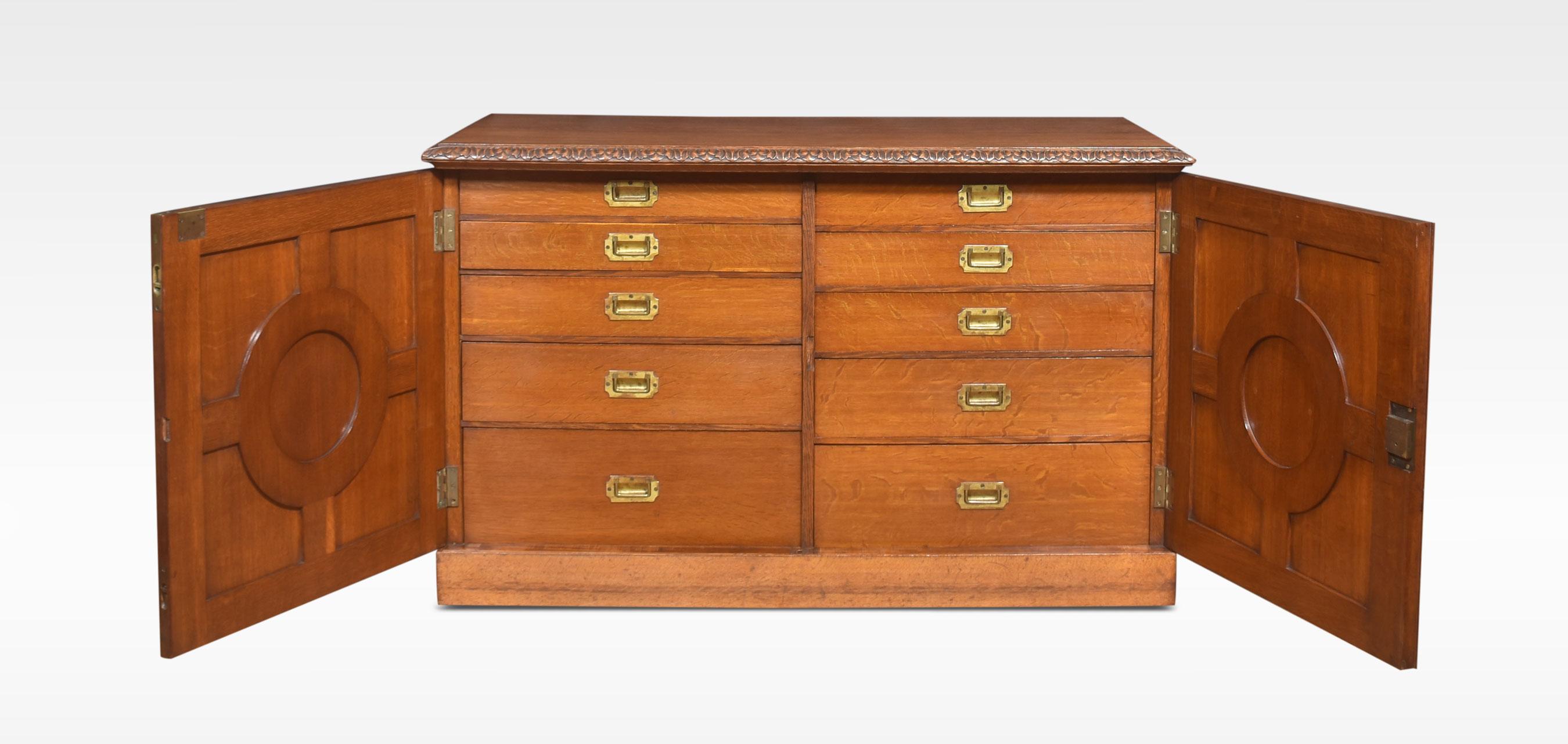 19th-century cabinet, the large rectangular well-figured oak top having a molded edge. Above two finely carved doors with foliated scrolling detail. The door opening to reveal two banks of five graduated drawers with recessed brass handles. All