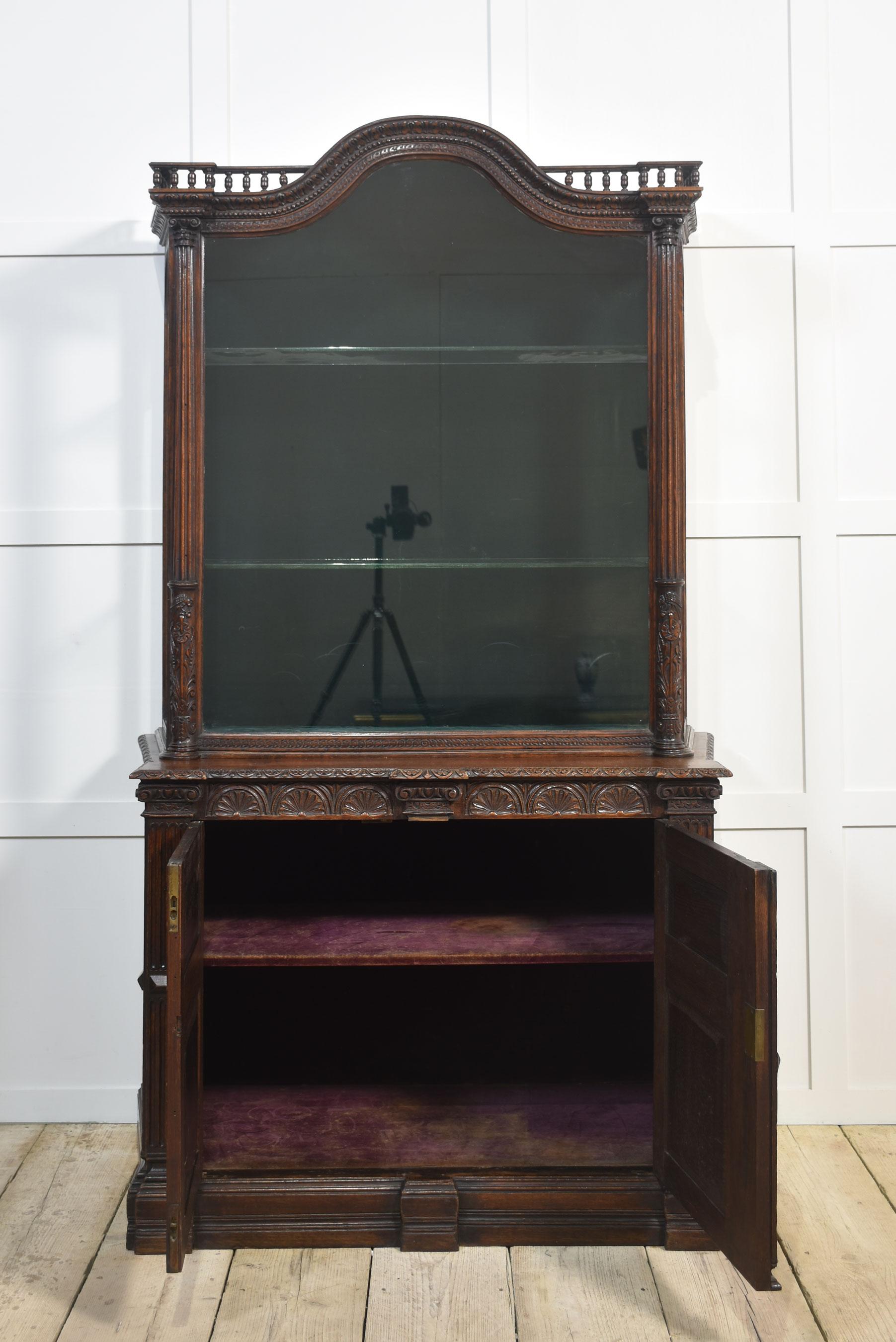 Oak display cabinet the shaped spindle top above large glazed door opening to reveal velvet lined interior and two glass shelves. To the heavily carved base section fitted with a pair of panelled doors with floral scrolling detail and reeded