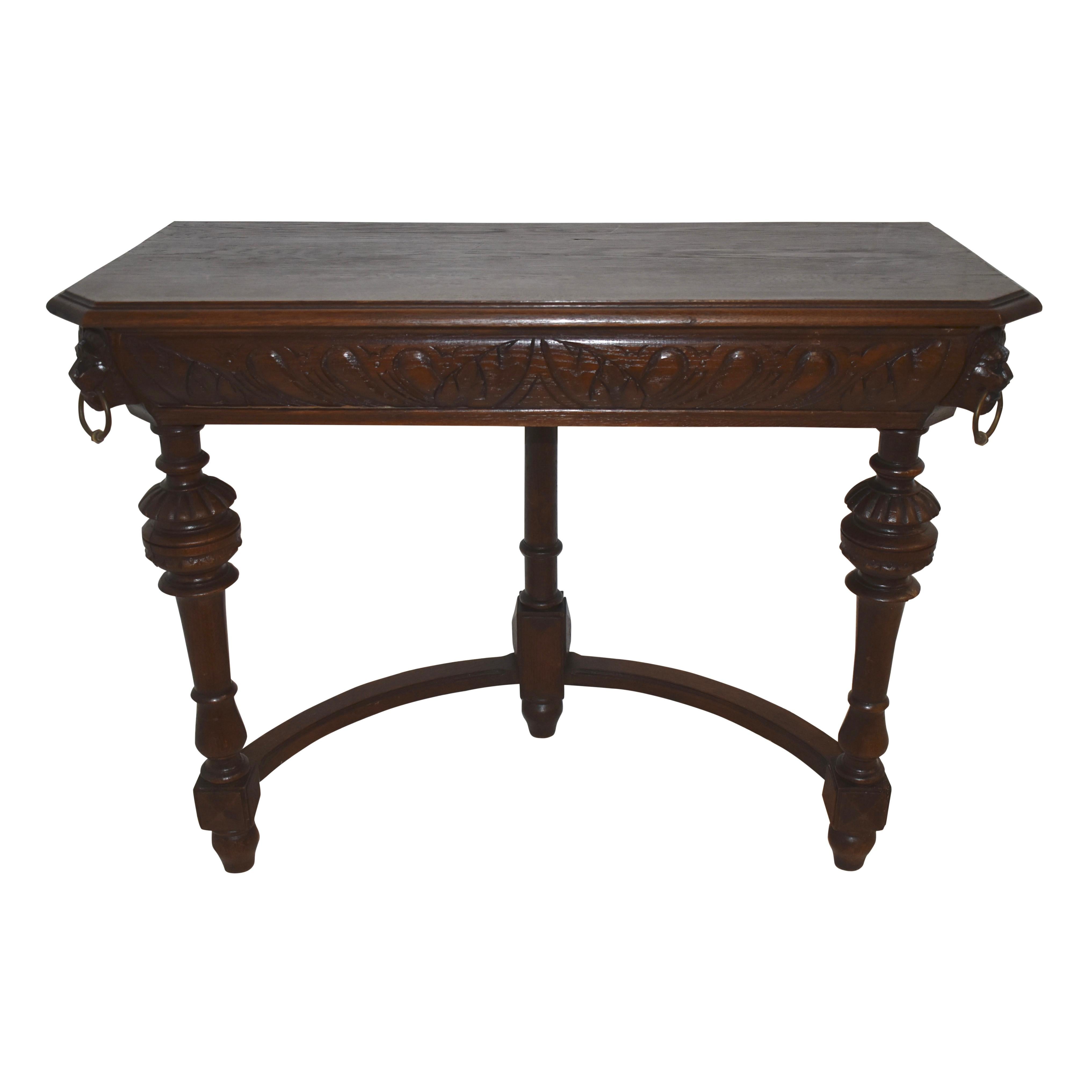 Supported by three turned legs united by a semicircle stretcher, this elegant hall table features carved lion heads with brass rings in their mouths at the table's canted corners. A beveled top, carved apron, and dark stain enhance the table's