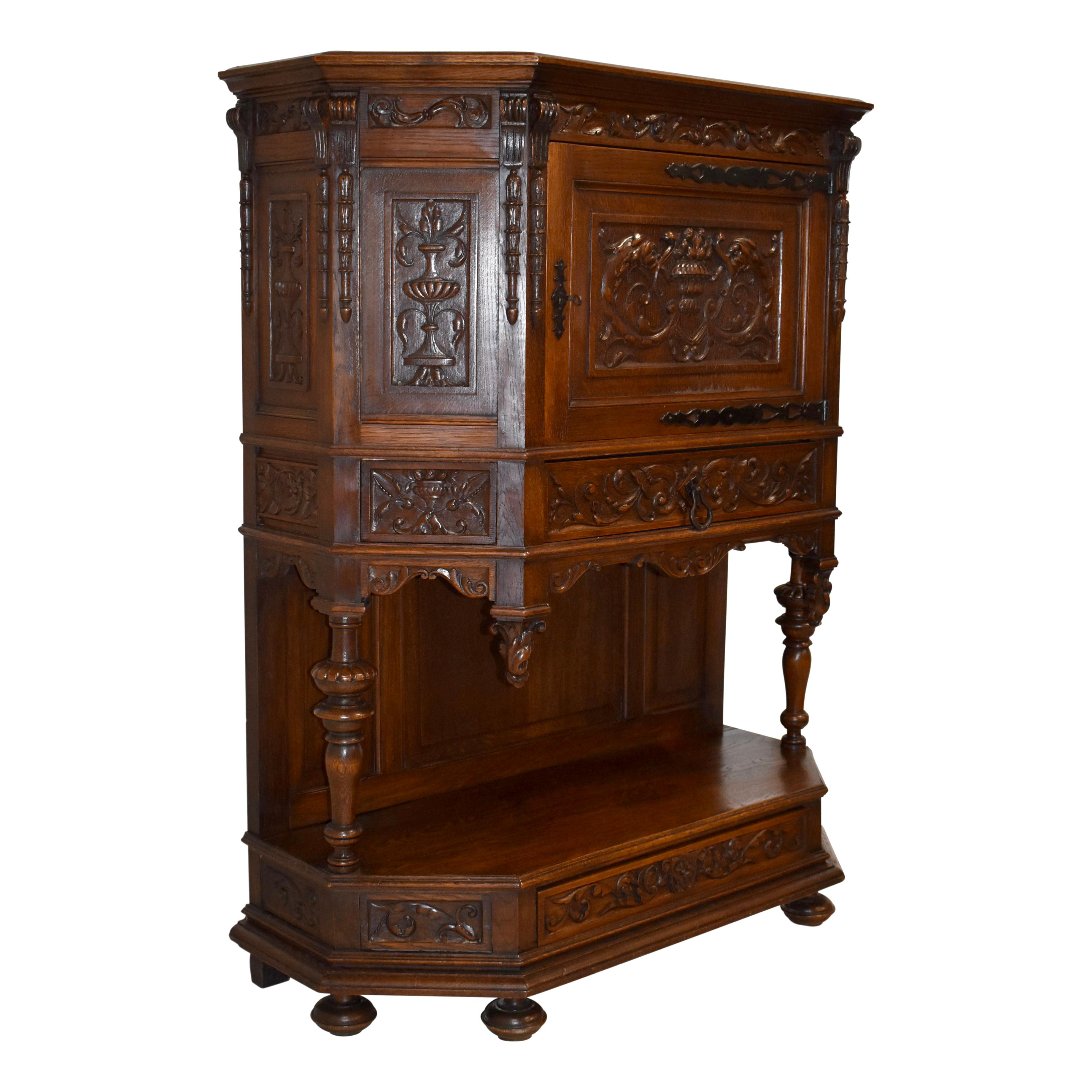 This heavily carved oak cabinet features a top with canted sides and a single door that showcases an urn flanked by opposing fish. The door swings on elegant, long hinges and opens to a single shelf. Below the door, a carved drawer provides