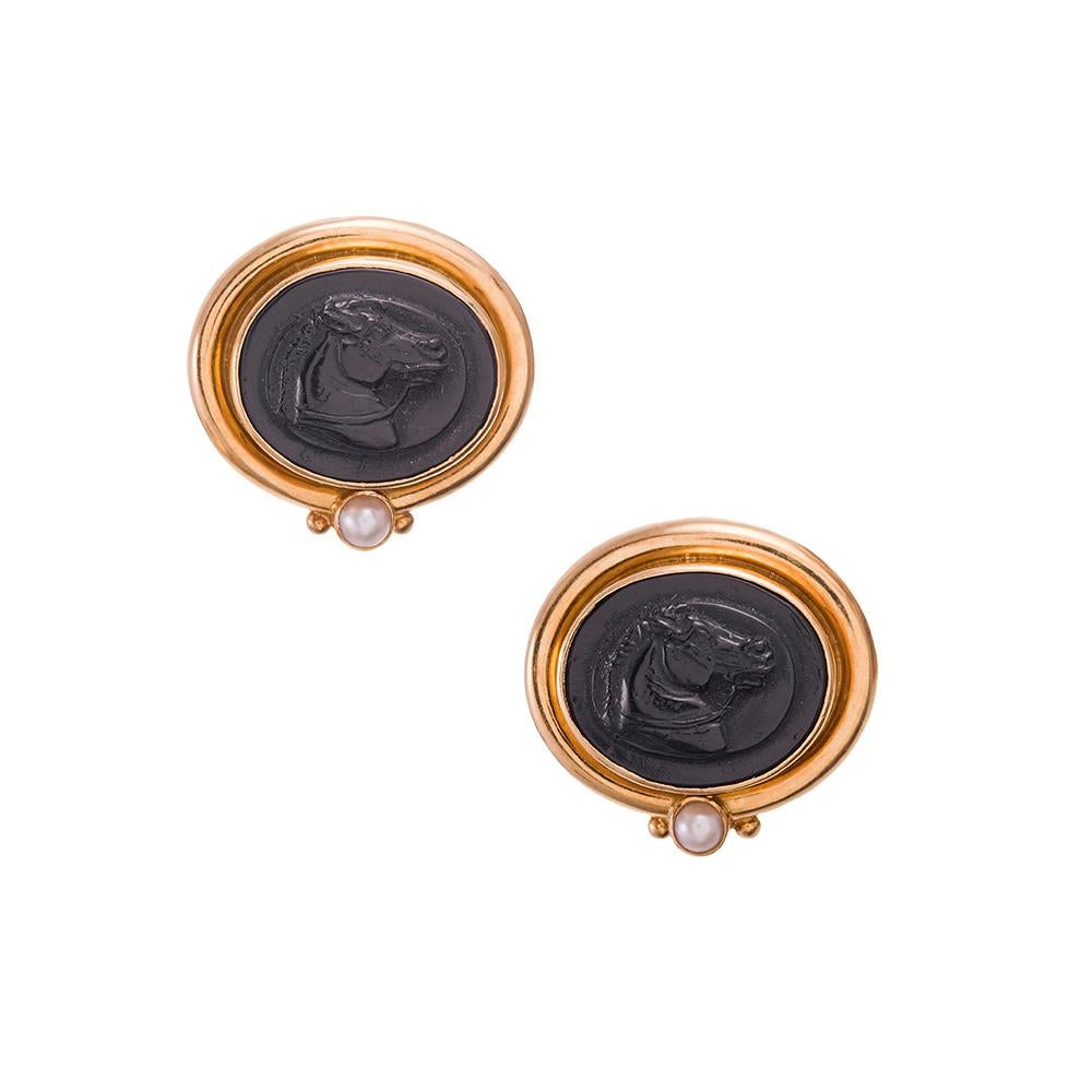 A pair of profile portraits of horses are immortalized in striking fashion, carved of black onyx and framed with 18k yellow gold. A pearl softens the design and adds a levity to the creation. Signed Elizabeth Locke. 1 inch by 7/8 of an inch.