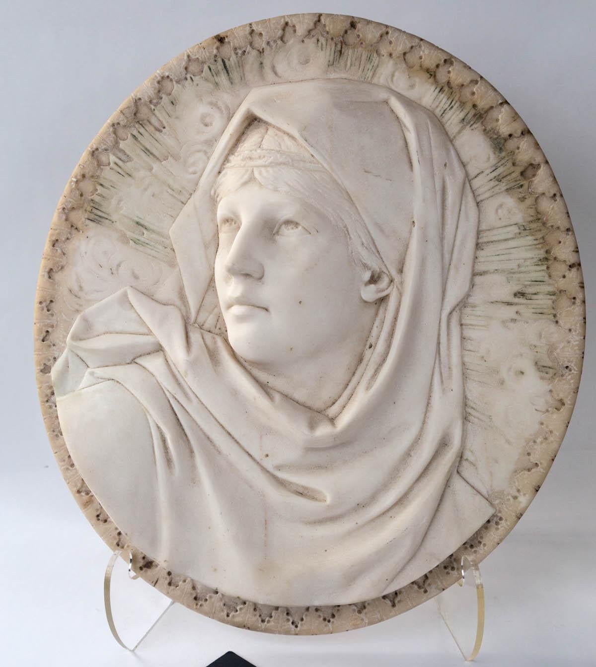 The Madonna wears a shawl around her head. Her eyes are cast upwards. Two tear drops on her left cheek. 
The marble is decorated around the subject with what seems to be rays of the sun. Ovoid swirls on the rays. The edges of the oval