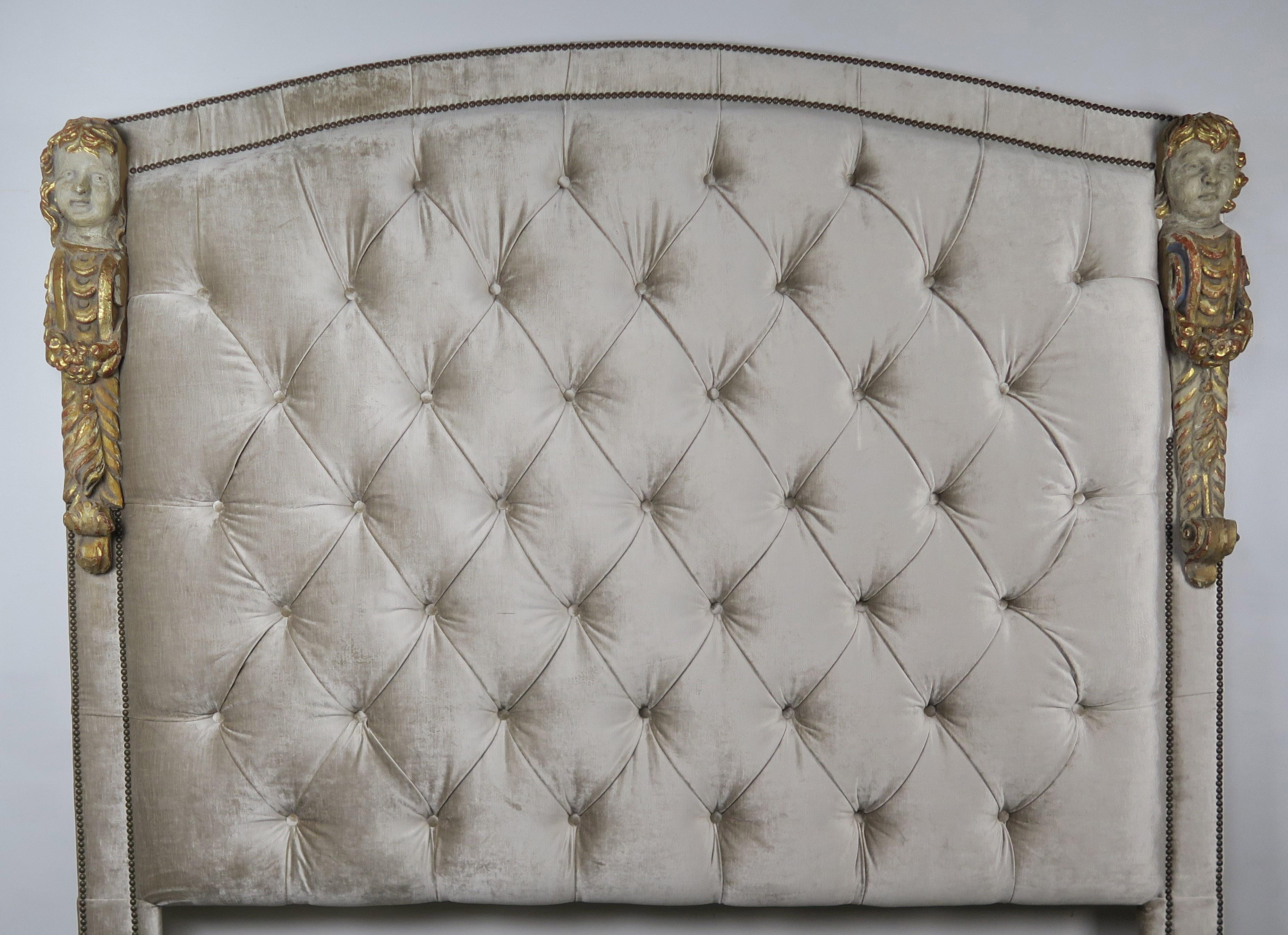 Custom headboard made with 19th century polychrome and giltwood cherubs. The 19th century figures are incorporated into a champagne colored velvet tufted headboard with a soft eye brow arched top. Two rows of antique brass nailhead trim detail