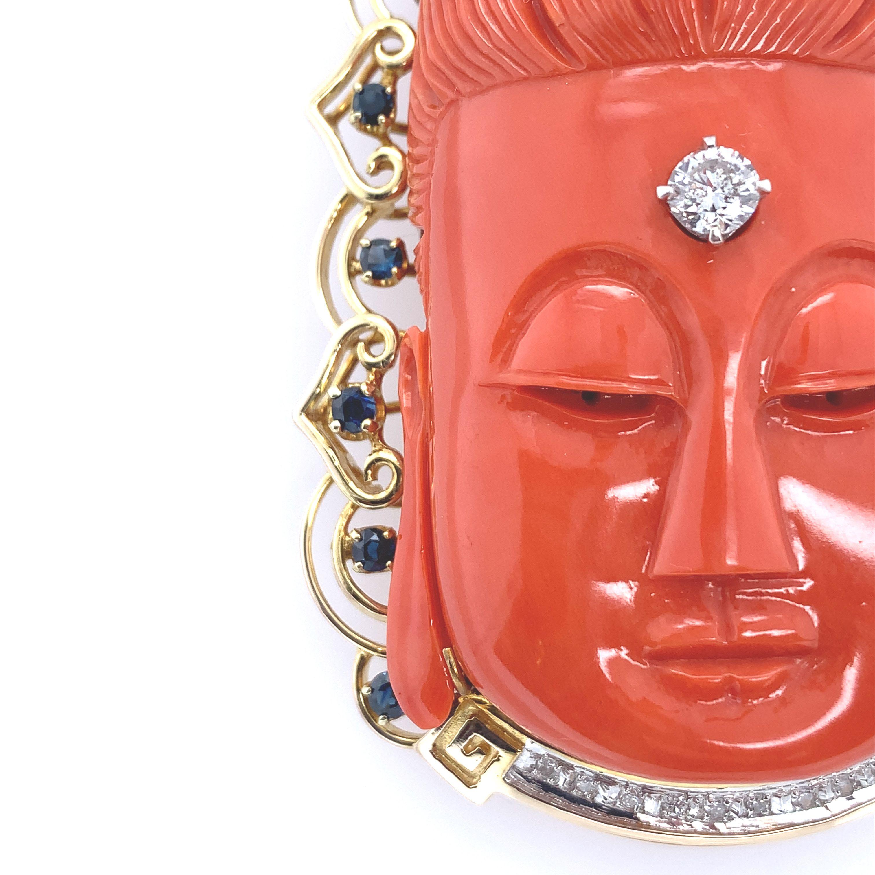 Magnificent large vintage Buddha pendant brooch set in 14K yellow gold. This intricate brooch features Buddha carved in natural red coral embraced by a decorative gold design featuring single-cut blue sapphires and diamonds. 

The coral carving