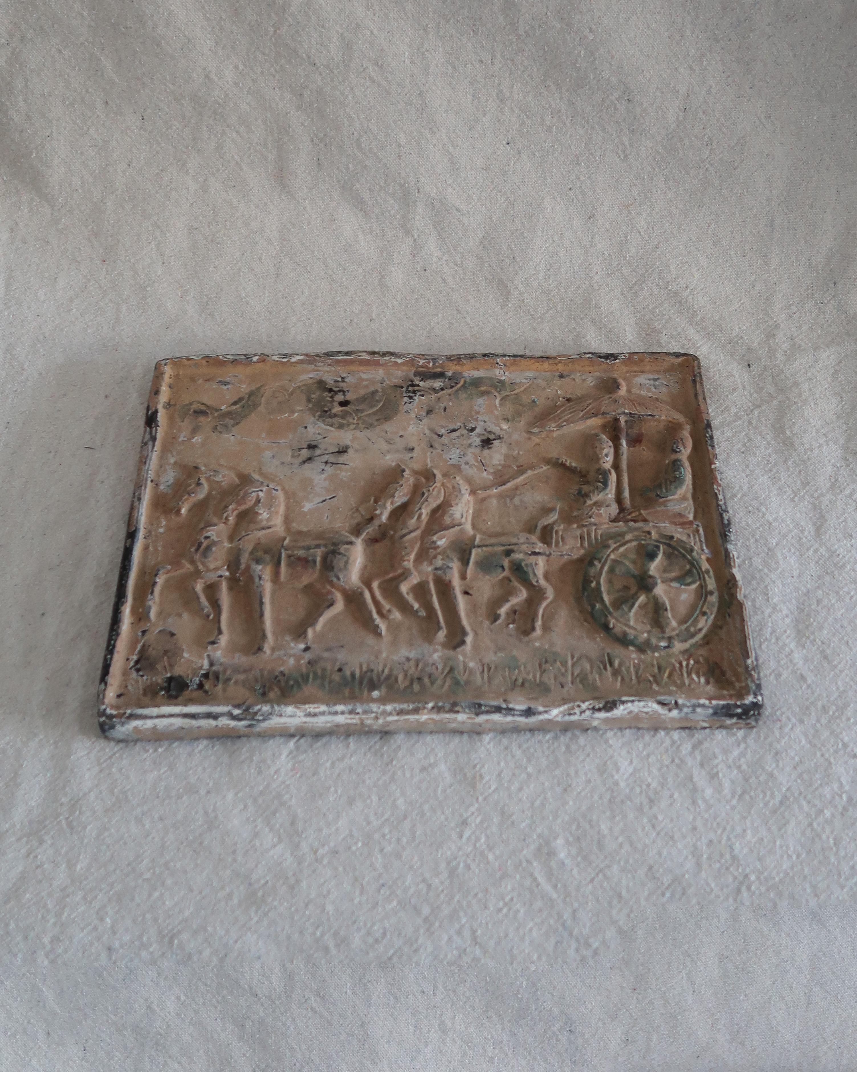 Une scène en bas-relief pleine de mouvement et de mystère. Cette plaque moulée représente un cortège de quatre chevaux tirant un char à roues avec deux personnages vêtus assis sous un parasol. La surface est usée et doucement peinte dans des tons de