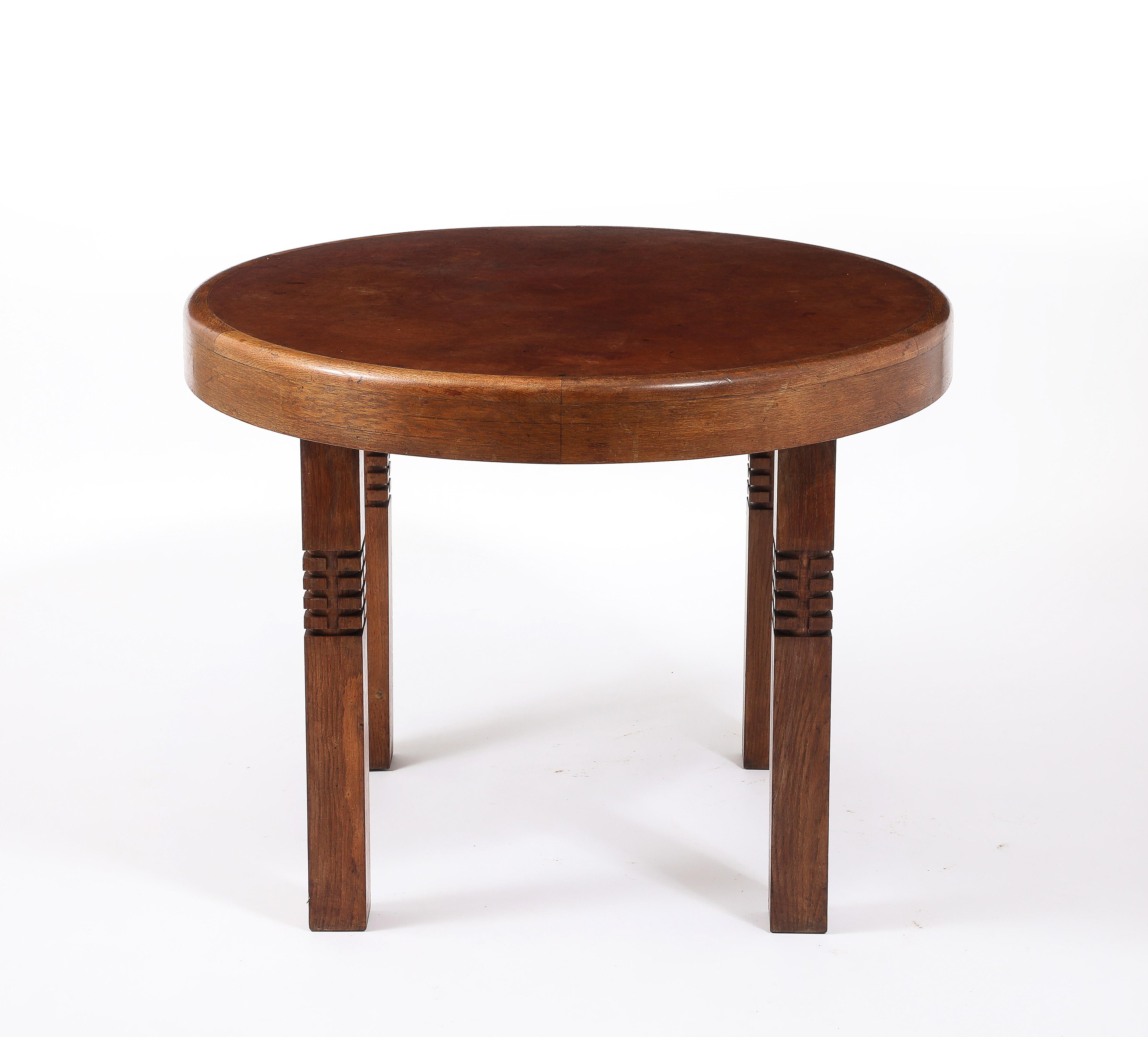 An elegant round center table with a rounded edge, the center is wood treated to have the appearance of leather. It is on four solid square legs, each carved with an intricate hidden round detail.