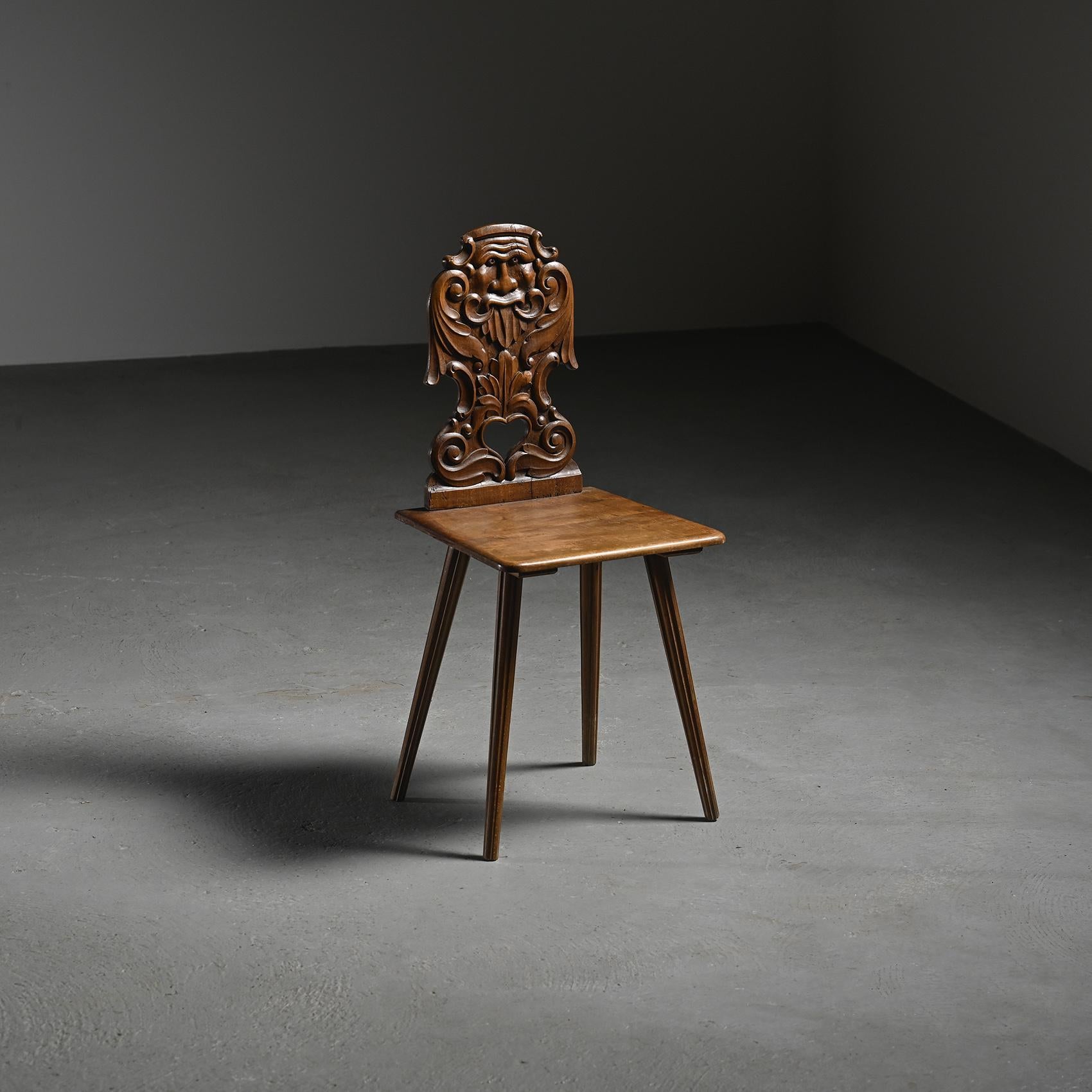 
Traditional escabelle chair with a carved backrest, made from solid turned wood, characteristic of Alpine folk art. 

The back panel is finely sculpted with a stylized face, or mascaron, framed by swirling vegetal forms, and features a heart-shaped